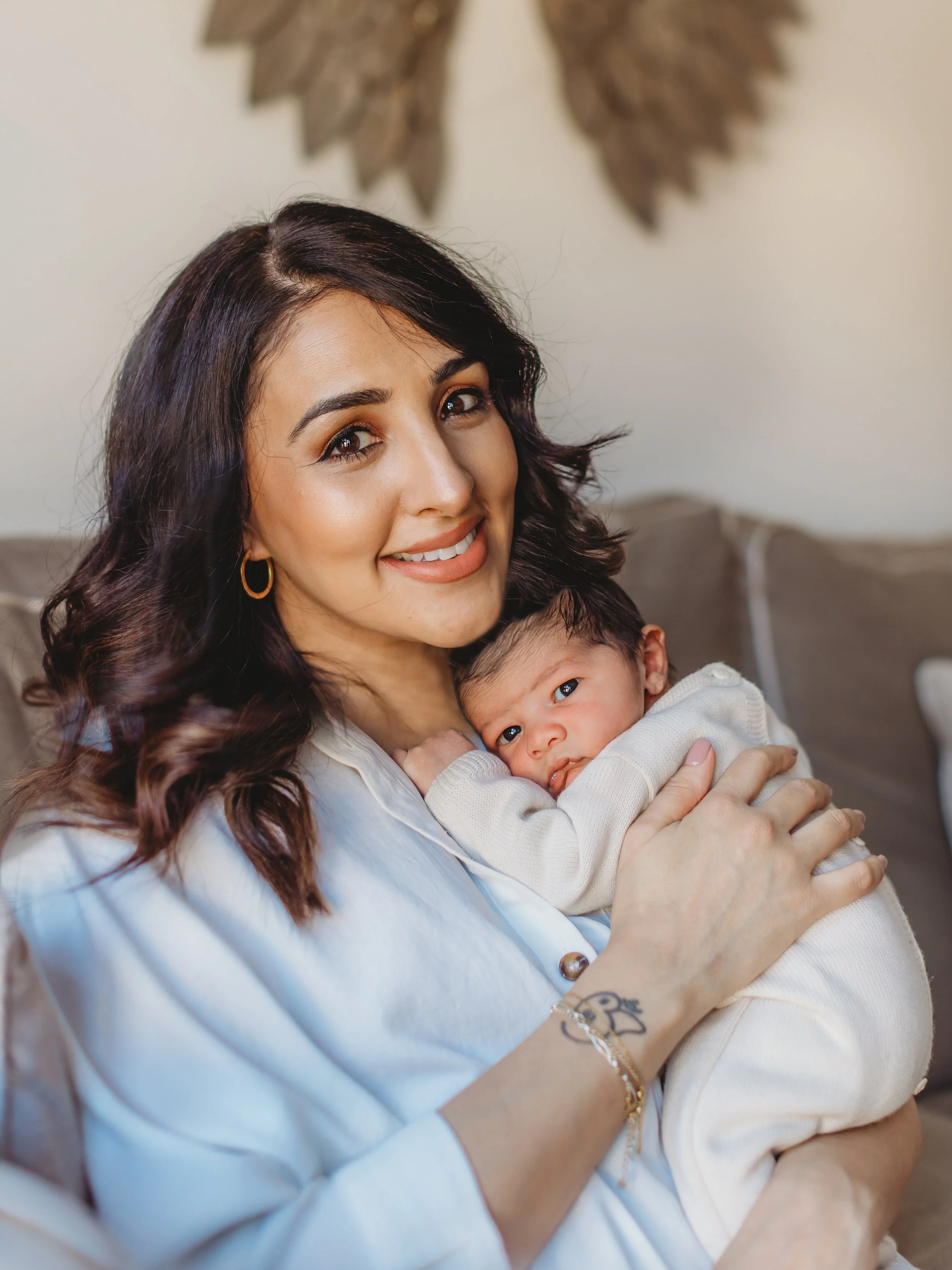 A woman with dark wavy hair and gold hoop earrings smiling while holding a newborn baby with dark hair and big eyes, sitting on a beige couch in a cozy living room.