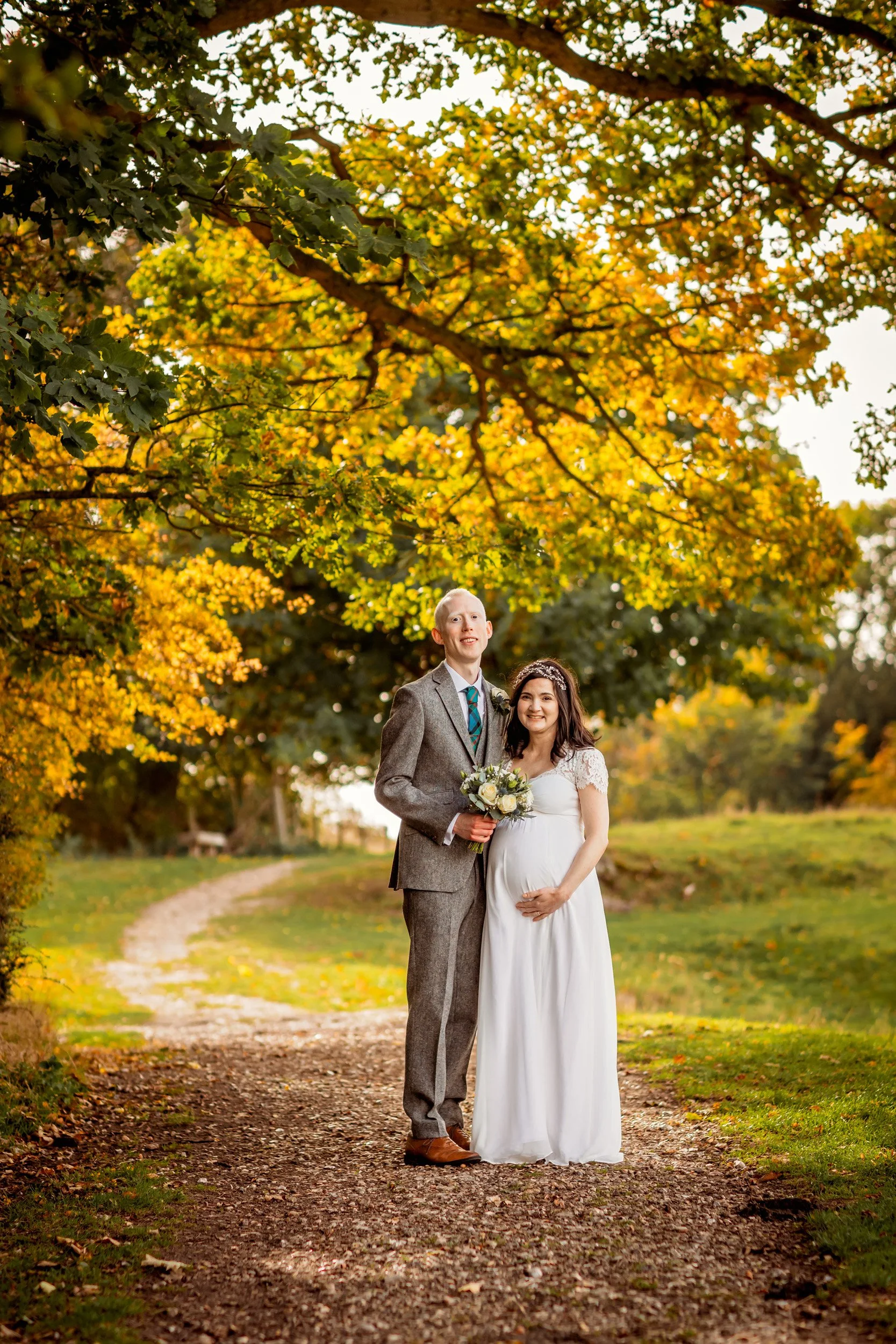 A pregnant woman in a white dress holding a bouquet and standing next to a man in a gray suit, on a leaf-covered path with autumn trees and yellow leaves overhead.