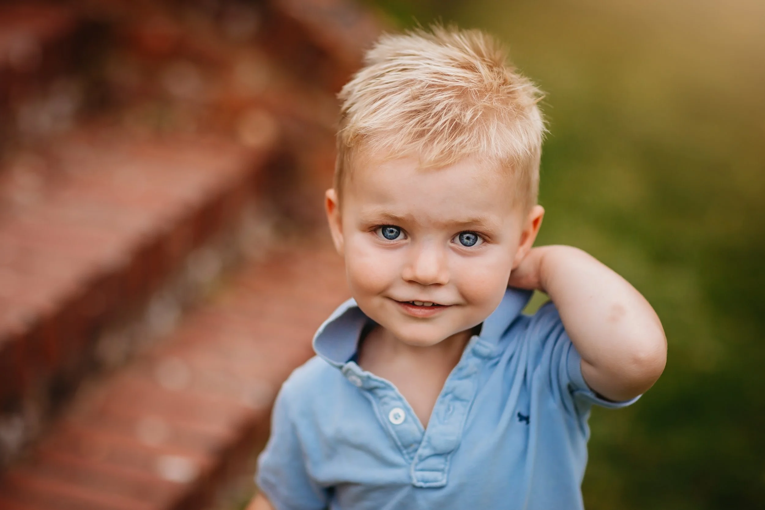 A young boy with blonde hair and blue eyes wearing a light blue polo shirt outdoors, smiling and touching the back of his neck.