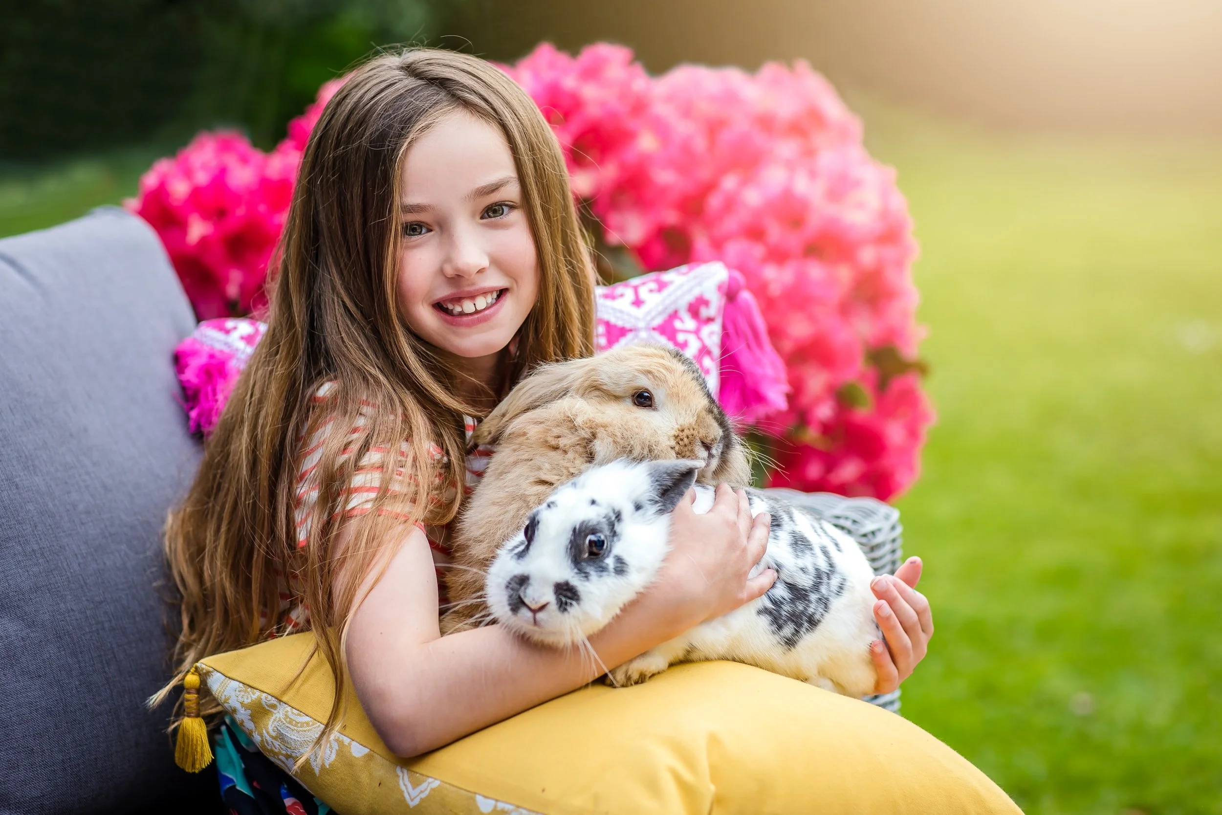 A young girl with long brown hair smiling and holding a rabbit, a guinea pig, and a rabbit with white and black fur, sitting outdoors on a yellow cushion with pink flowers in the background.
