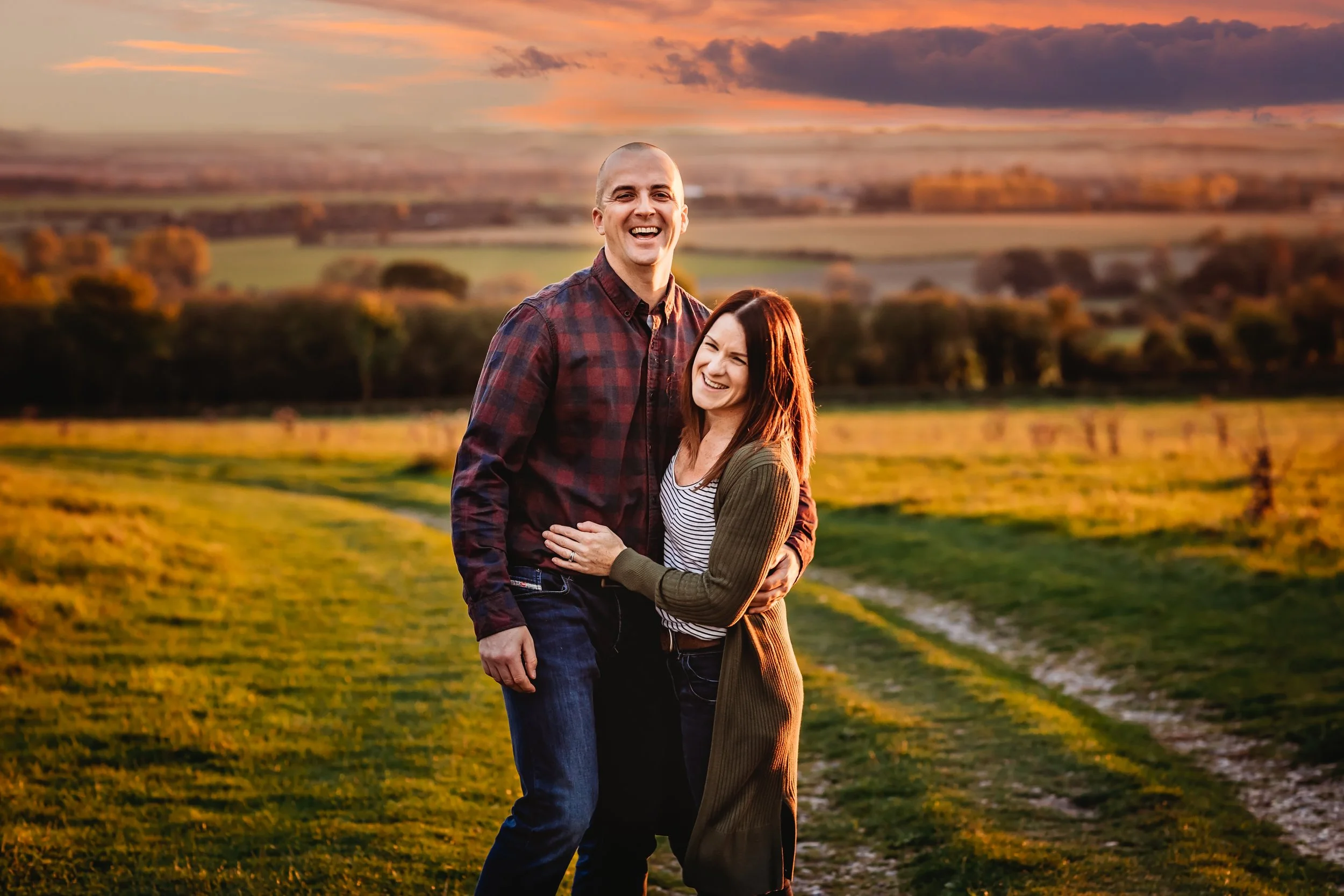A happy couple standing close together on a countryside path during sunset, smiling and embracing with a scenic landscape in the background.