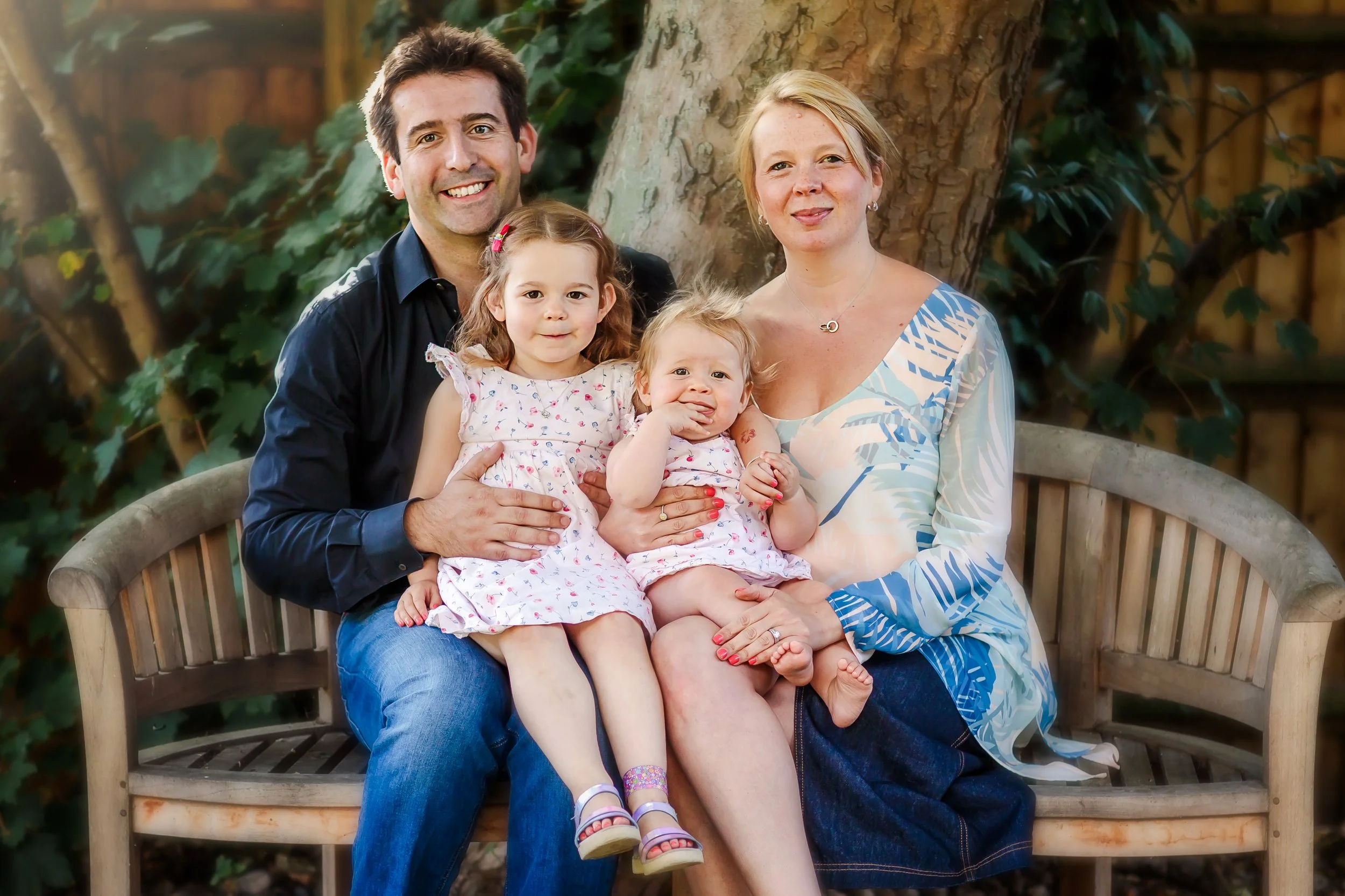 A family of four sitting on a wooden bench outdoors, with an adult man, an adult woman, and two young girls, all smiling at the camera.