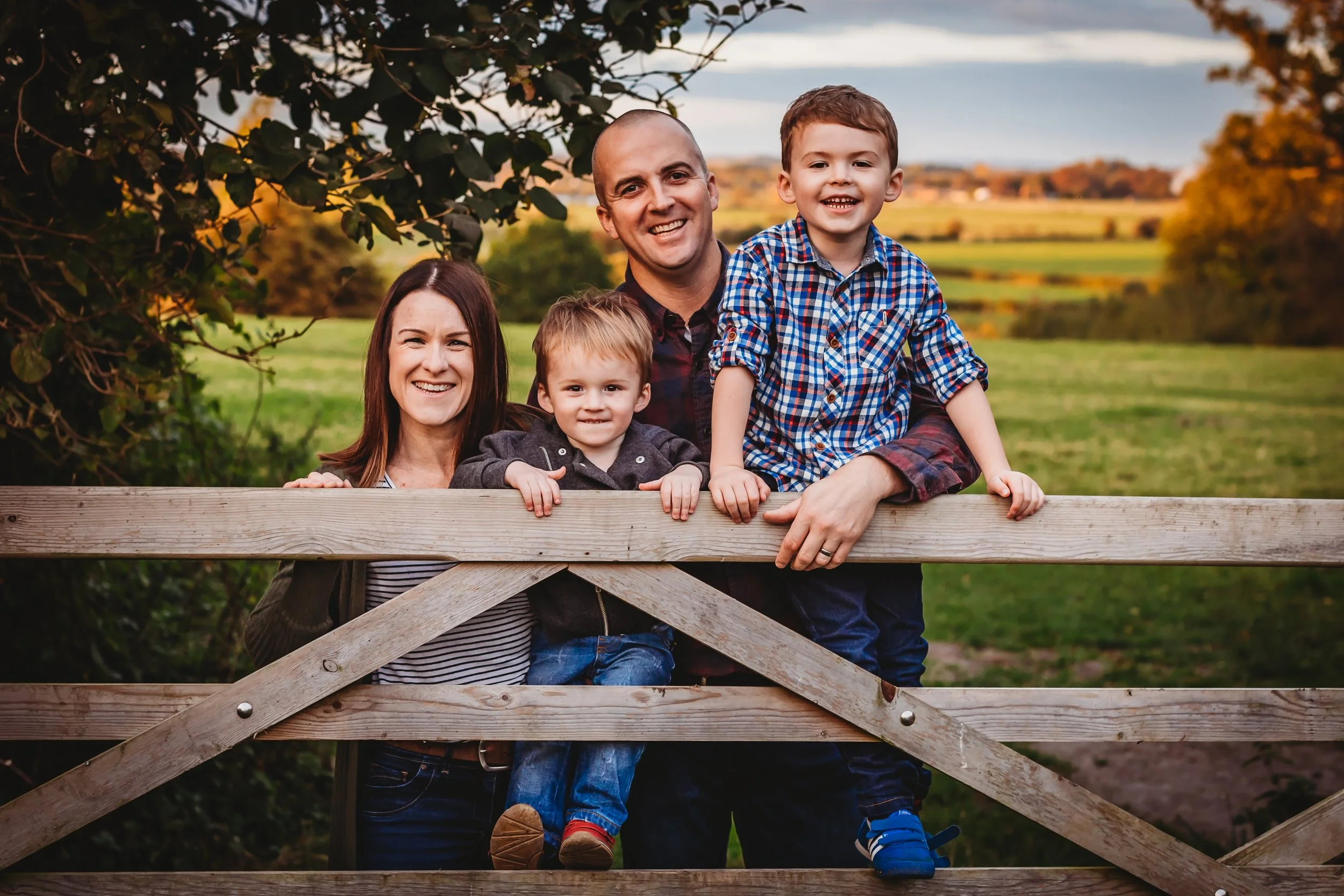 A smiling family of four, including a mother, father, and two young sons, stands behind a wooden fence outdoors, with trees and fields in the background during sunset.