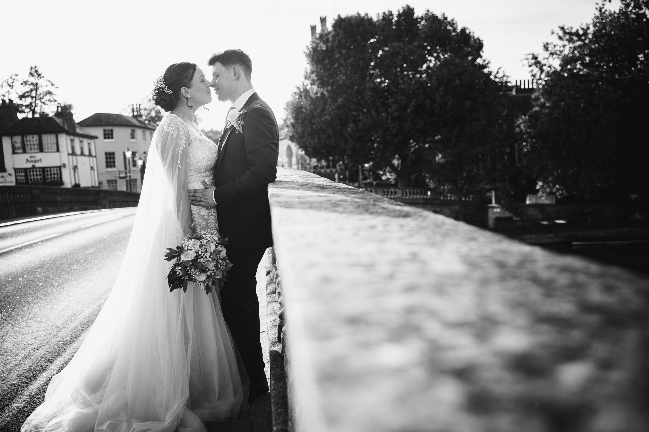 Black and white photo of a bride and groom standing closely on a bridge, facing each other with noses touching, holding a bouquet of flowers, with trees and buildings in the background.