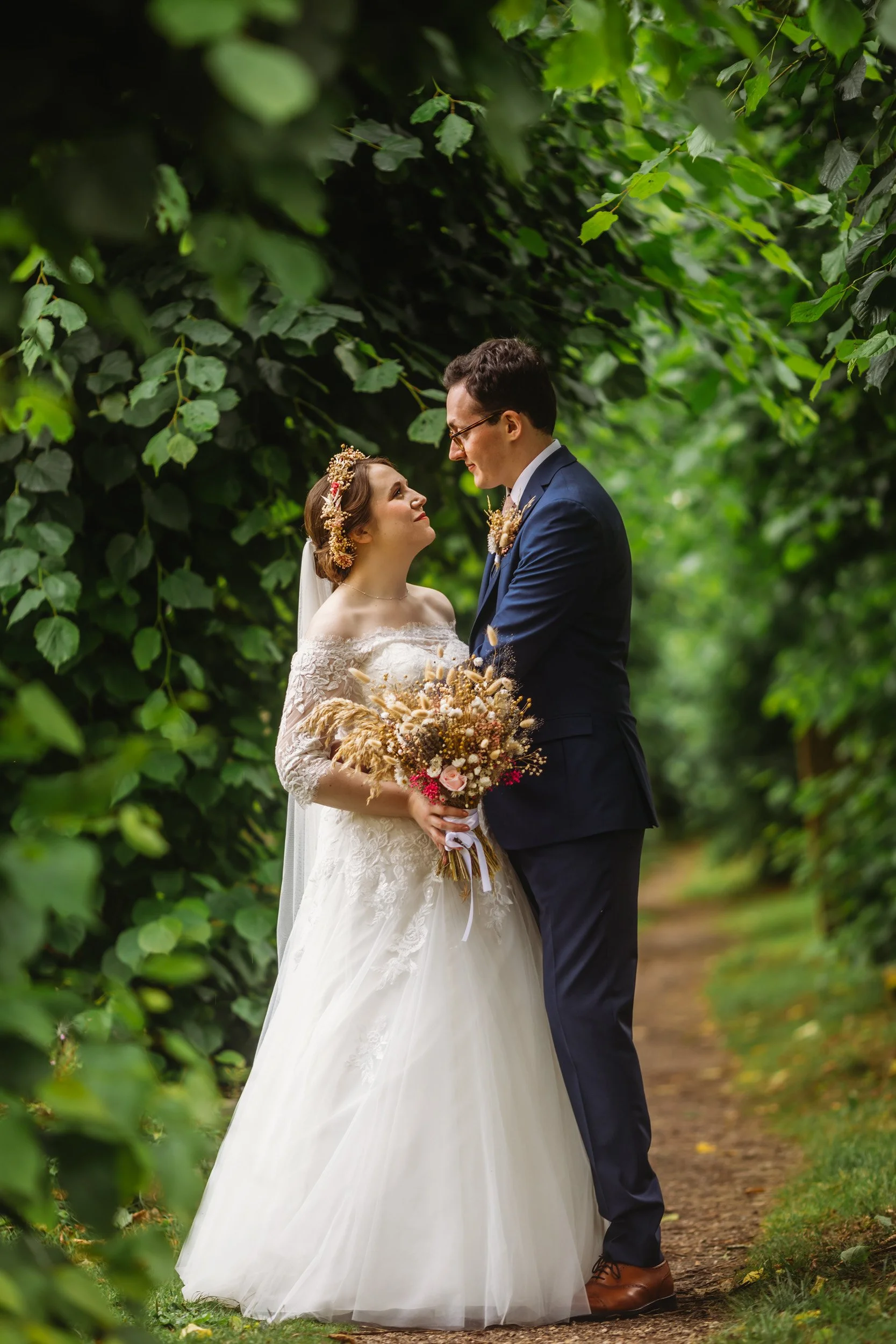 A bride and groom on their wedding day standing in a lush green outdoor setting, gazing into each other's eyes with their foreheads almost touching. The bride is wearing a white off-shoulder wedding gown and holding a bouquet of flowers, while the gr