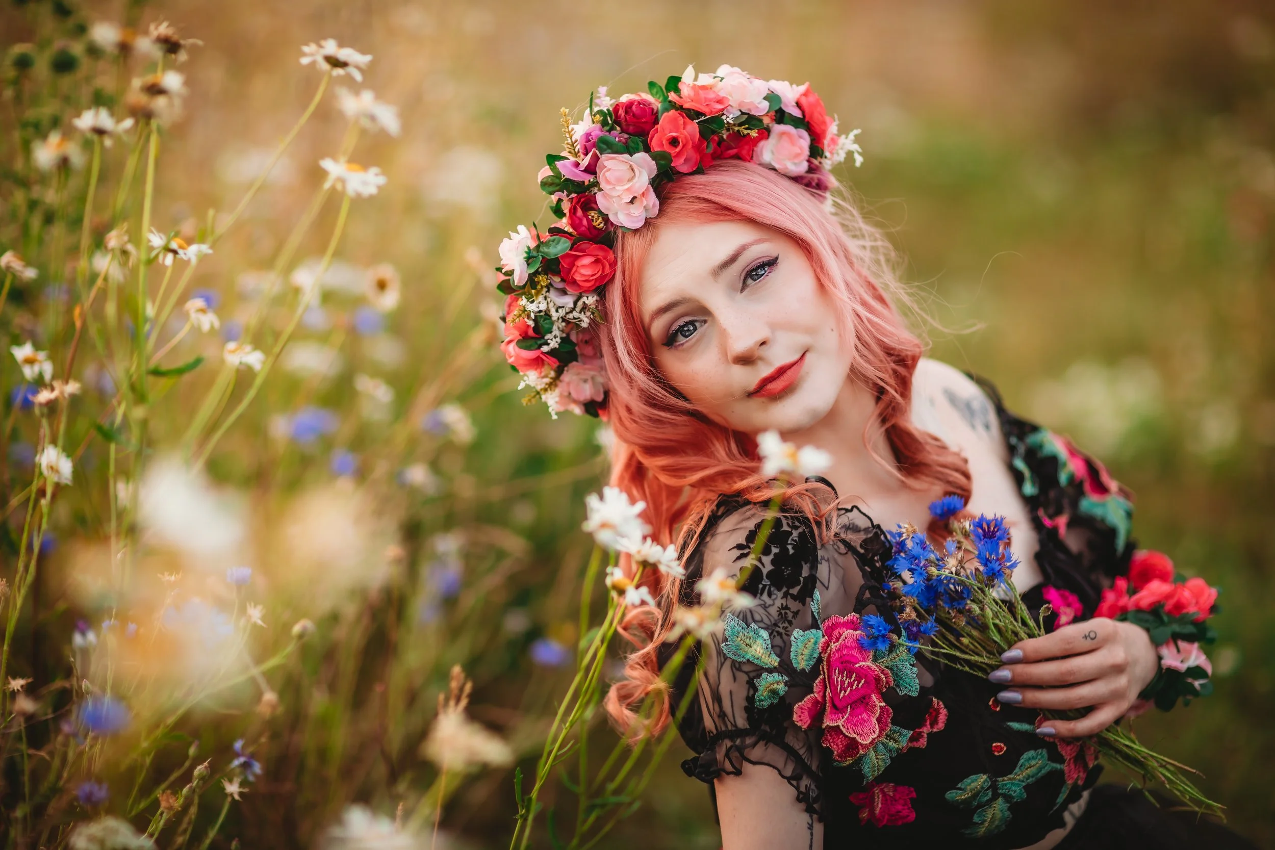 A woman with pink hair wearing a floral crown and a black embroidered dress holding a bouquet of flowers in a field of wildflowers.