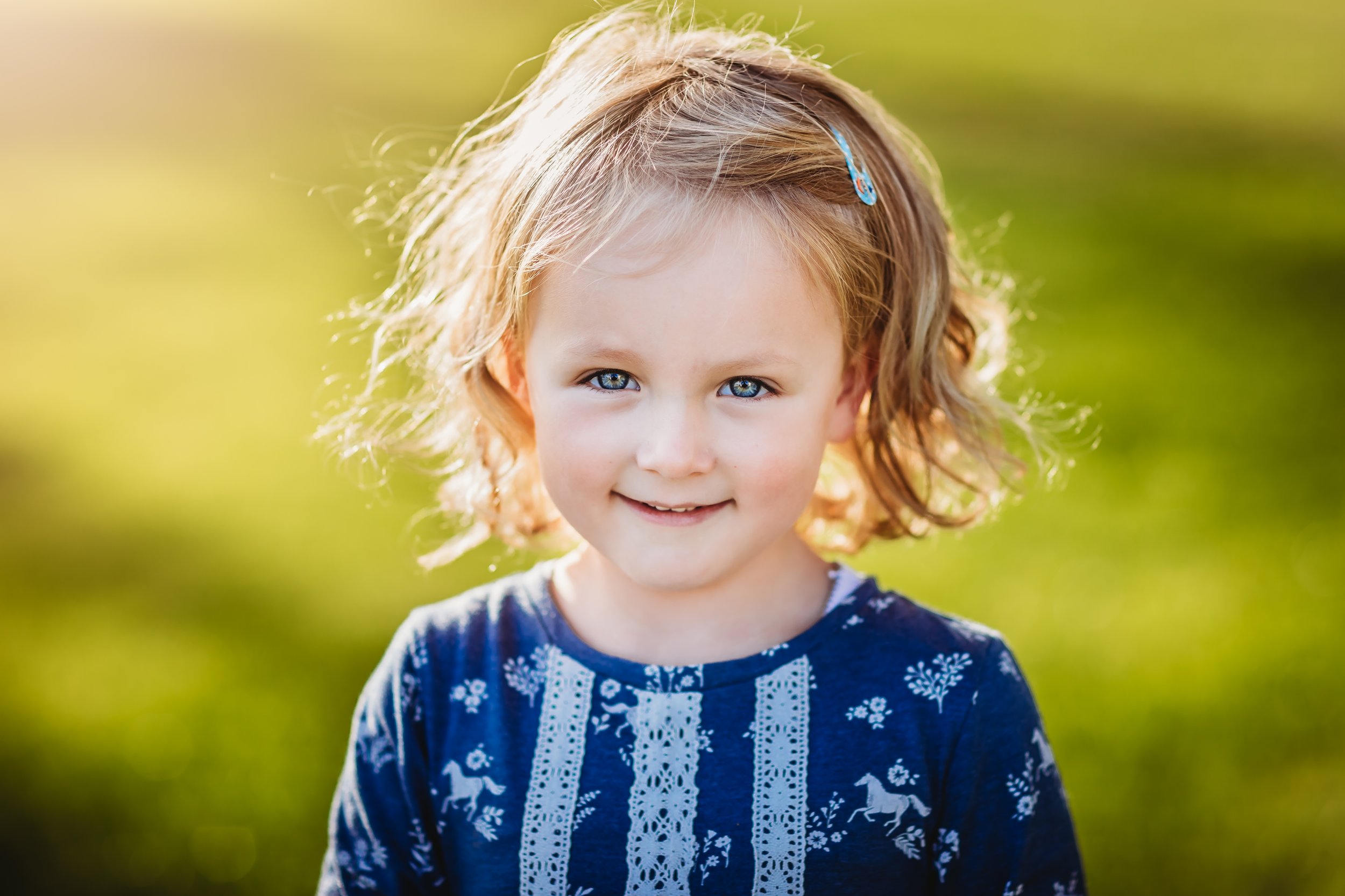 Close-up of a young girl with blond hair and blue eyes, smiling outdoors with blurred green background.