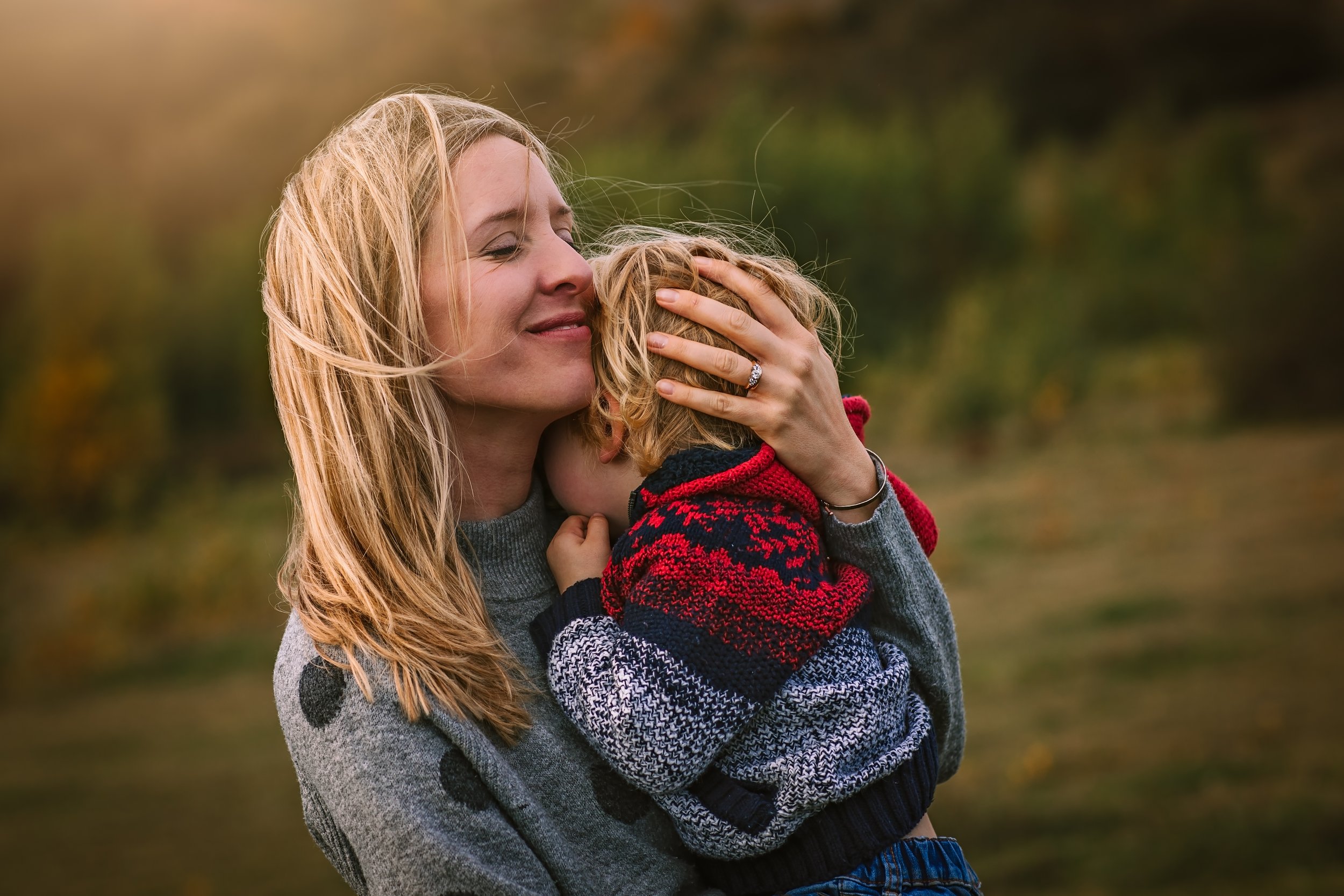 A woman with blonde hair holding and hugging a young child outdoors during autumn, with trees in the background.