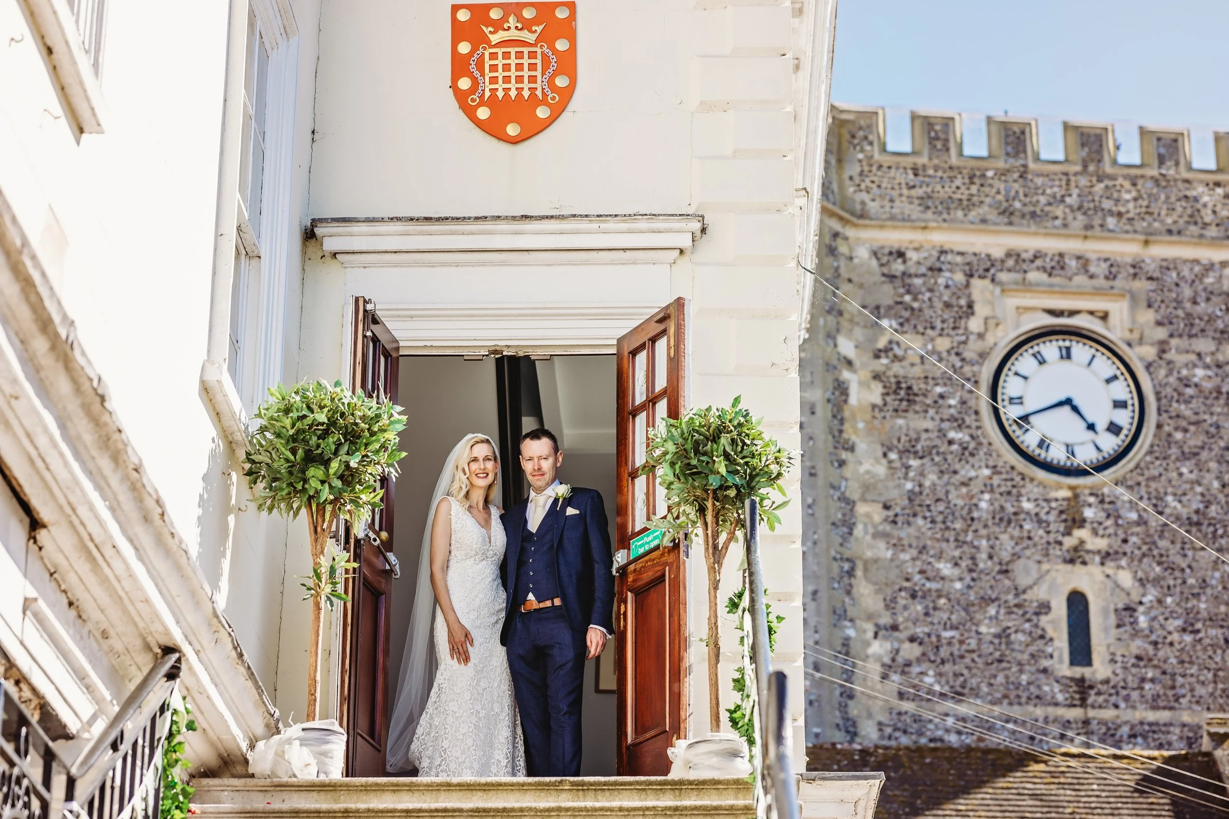 A bride and groom standing at the open doorway of a historic building during a wedding, with a clock tower in the background.