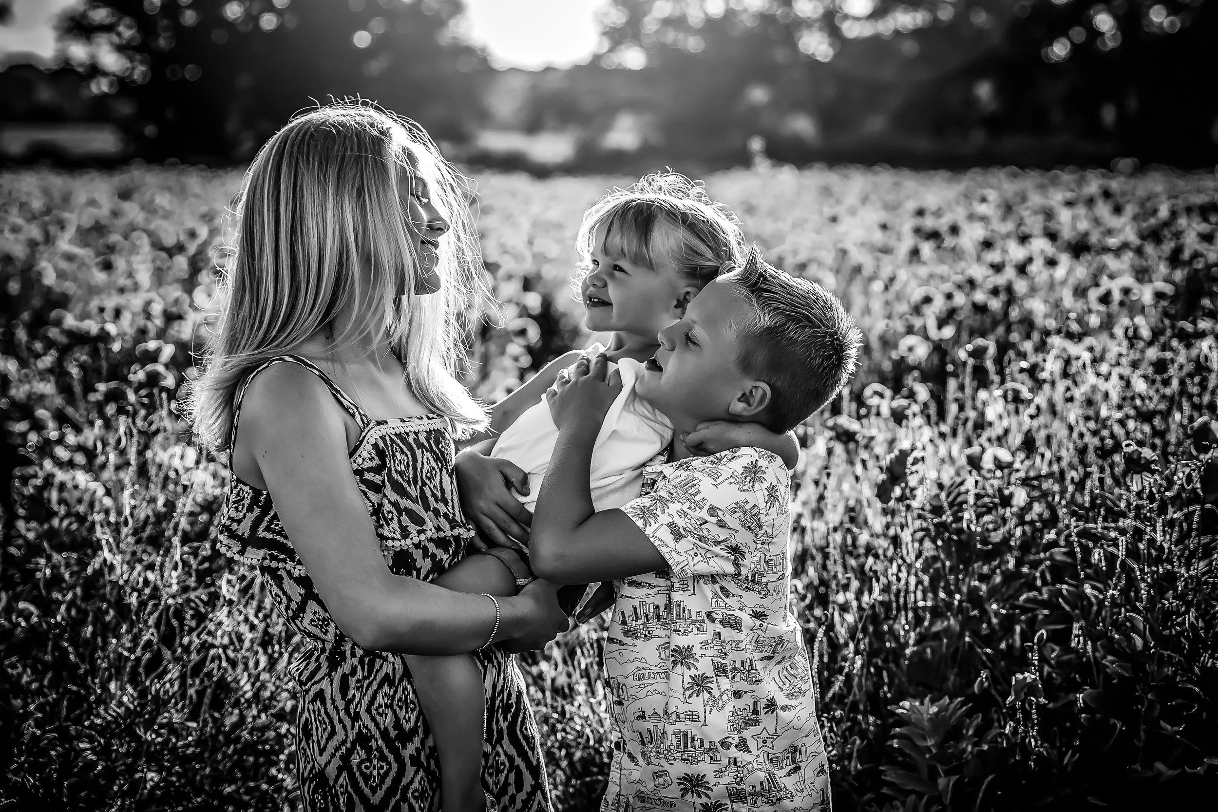 A woman holding a young girl in her arms while a boy embraces her from the front outdoors in a field, all smiling and looking at each other in black and white.