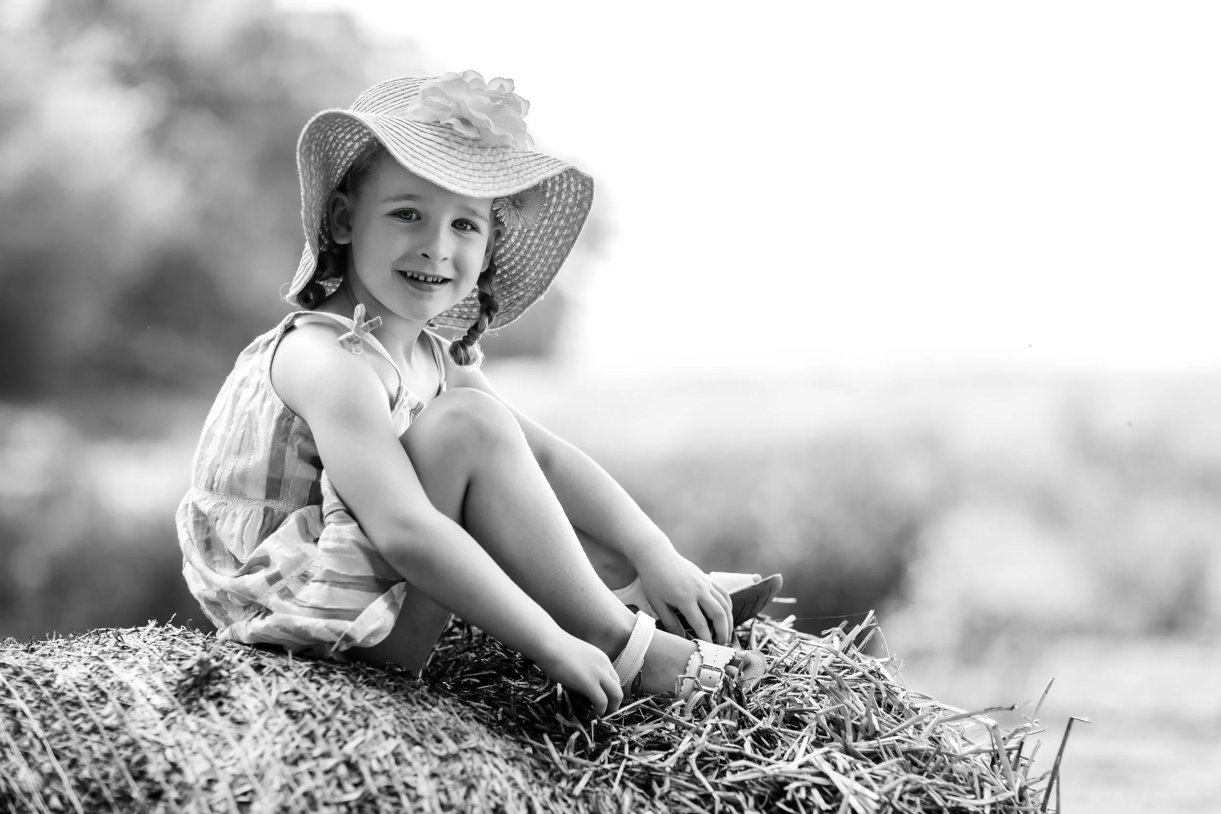A young girl sitting on a haystack outdoors, smiling at the camera, wearing a hat and a summer dress.