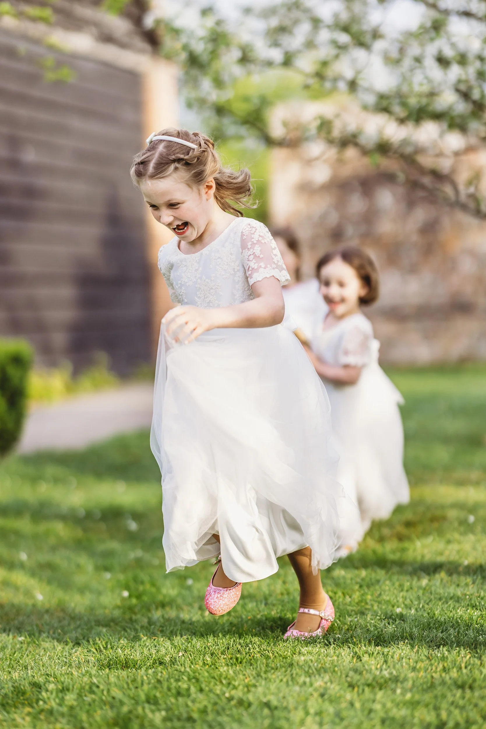 Children in white dresses playing outside on a grassy lawn, smiling and running in a joyful manner.