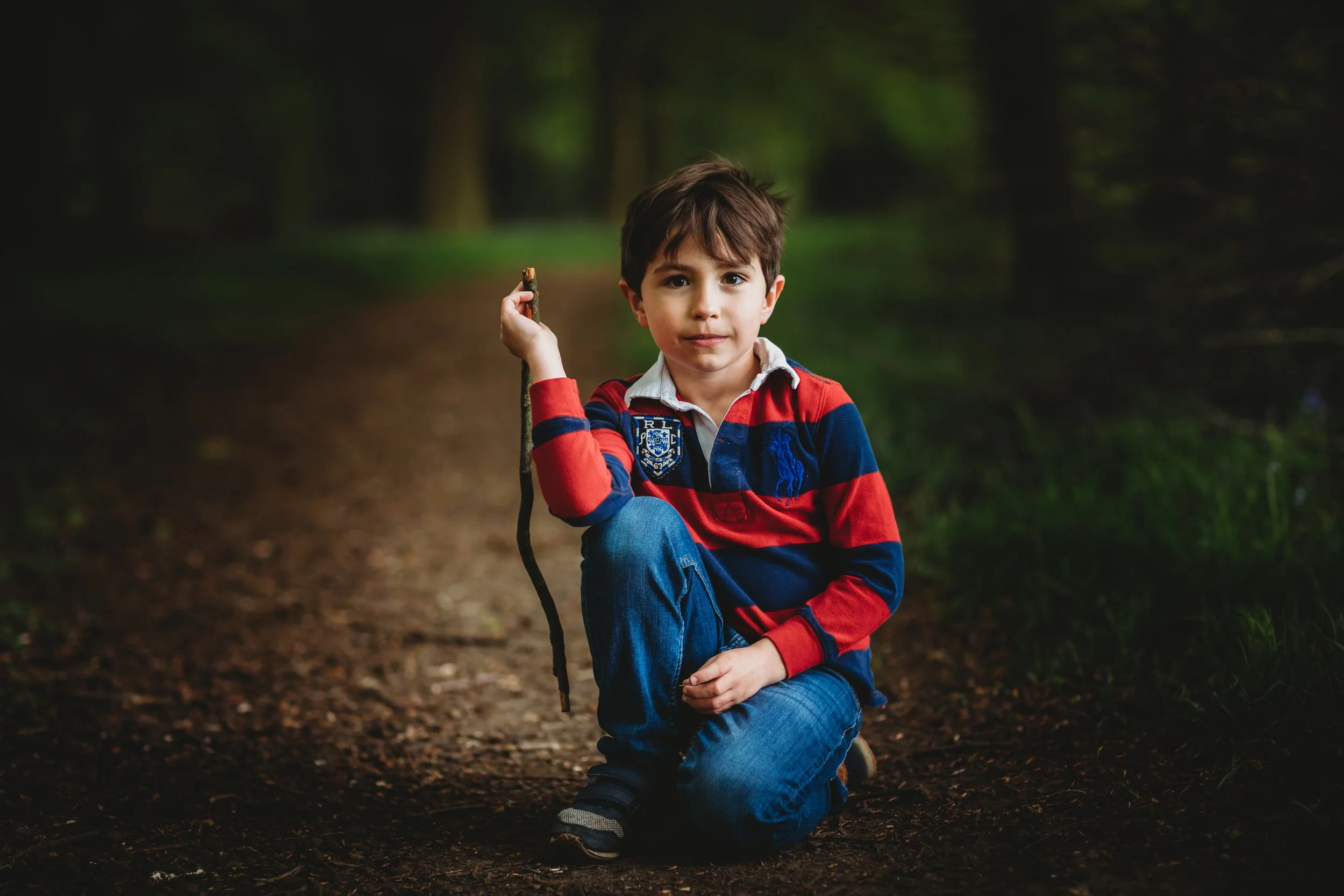 Young boy kneeling on a dirt path in a forest, holding a stick in his right hand, wearing a red and navy striped rugby shirt and jeans.
