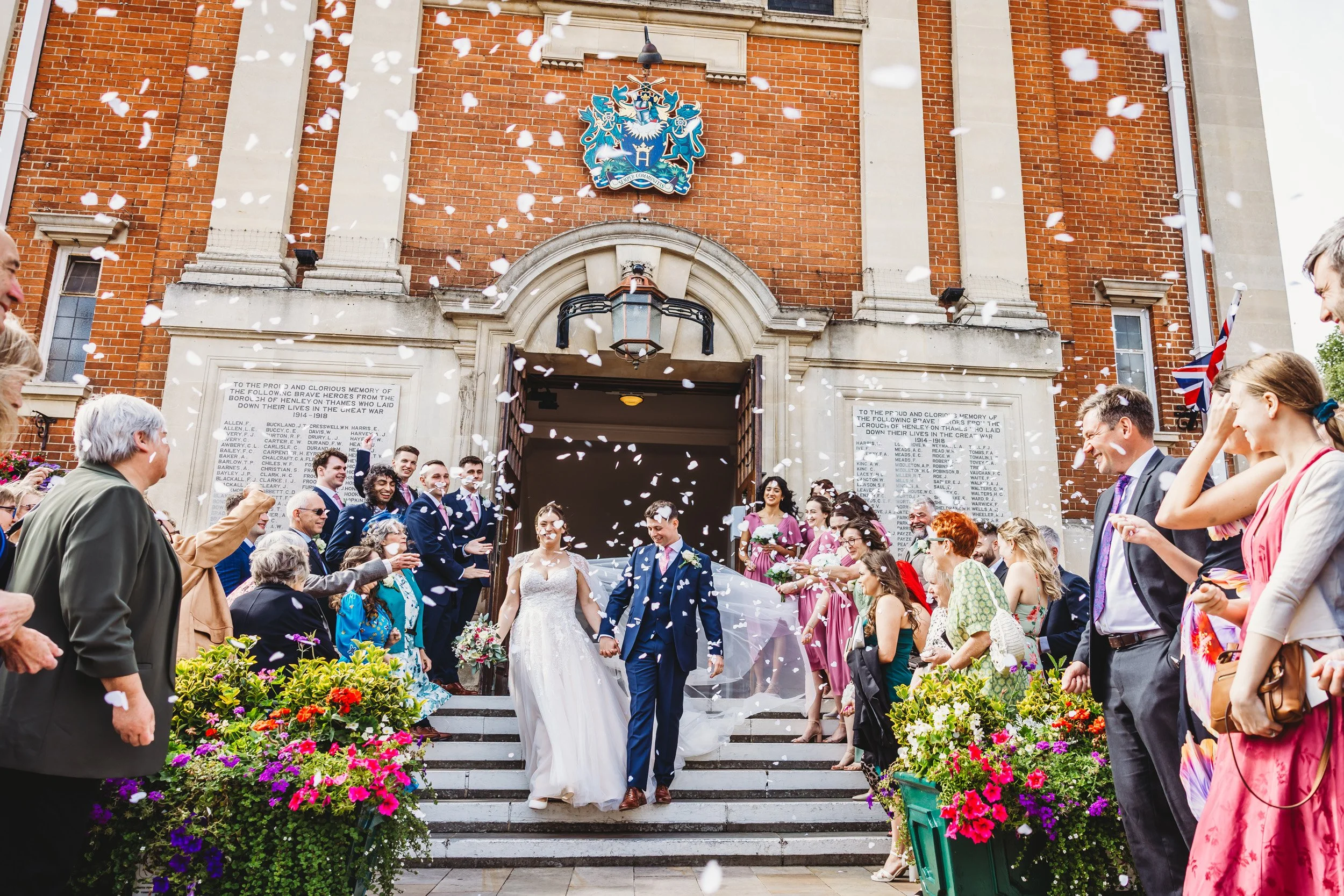 A wedding celebration with a bride and groom exiting a church, surrounded by guests throwing confetti and confetti falling in the air, with flowers decorating the steps and a coat of arms above the entrance.