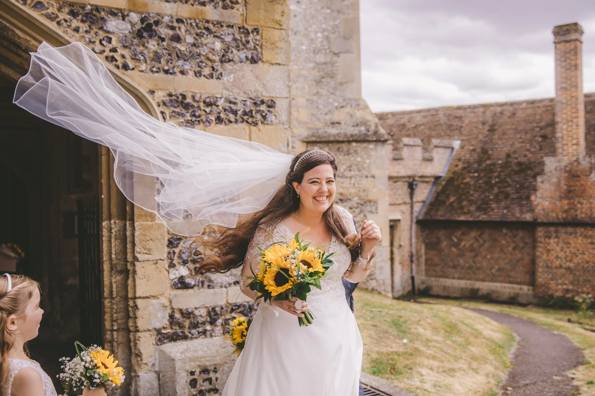 A smiling bride in a white wedding dress holding a bouquet of yellow sunflowers and white flowers, with her veil flowing in the wind, outside a stone church building, accompanied by a young flower girl holding a smaller bouquet of sunflowers.