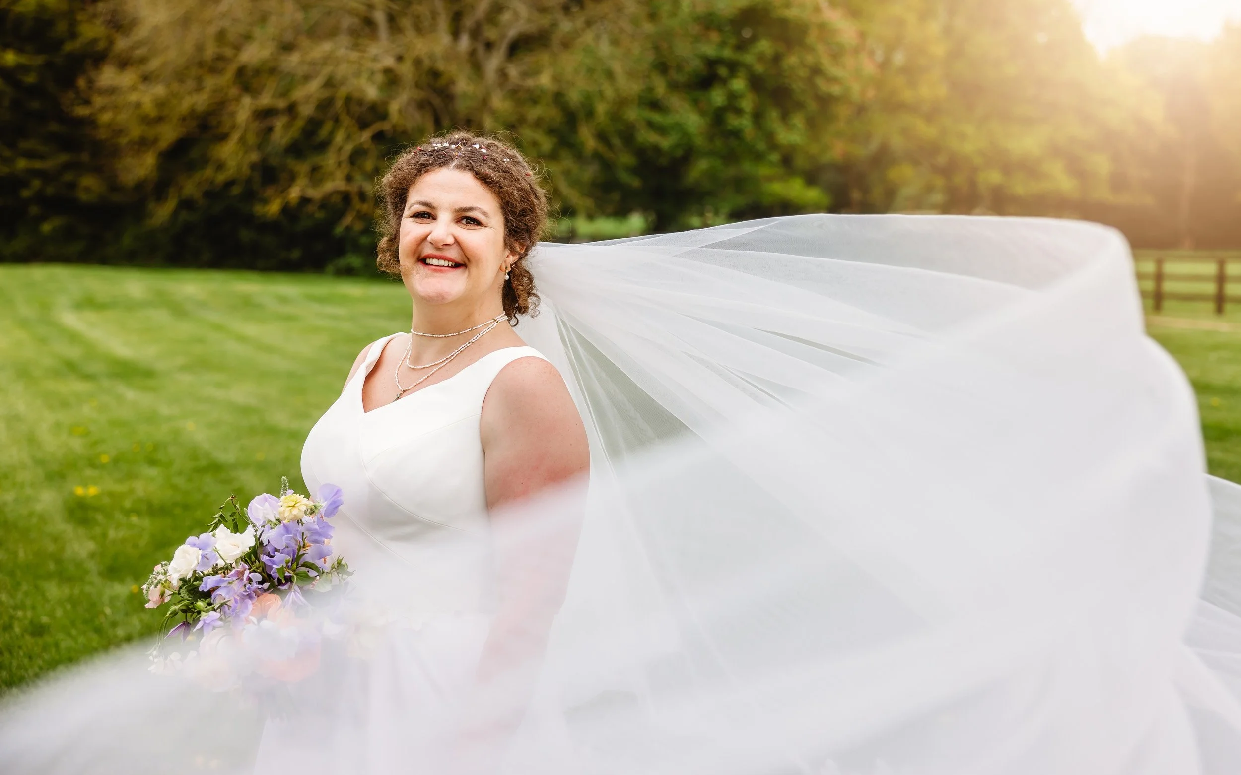 A smiling bride in a white wedding dress holding a bouquet of purple and white flowers outdoors, with a flowing veil and a green field in the background.