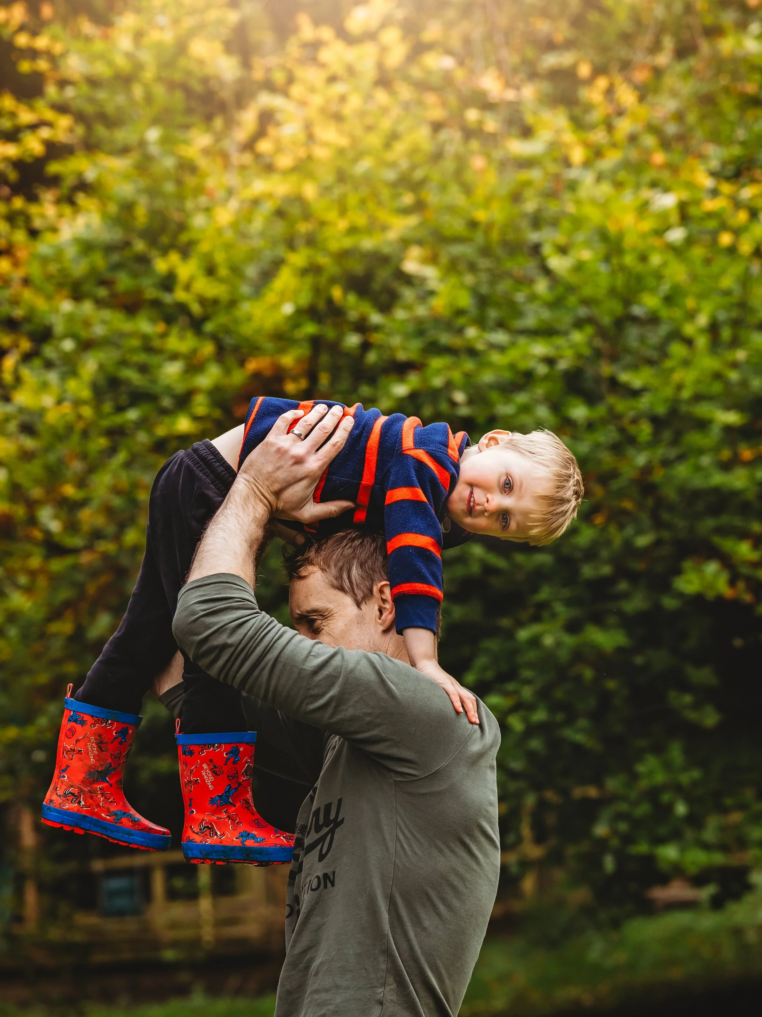 A man lifting a young boy with blonde hair in the air outdoors during autumn, with trees showing fall foliage in the background. The boy is wearing colorful rain boots and a navy blue sweater with red stripes.