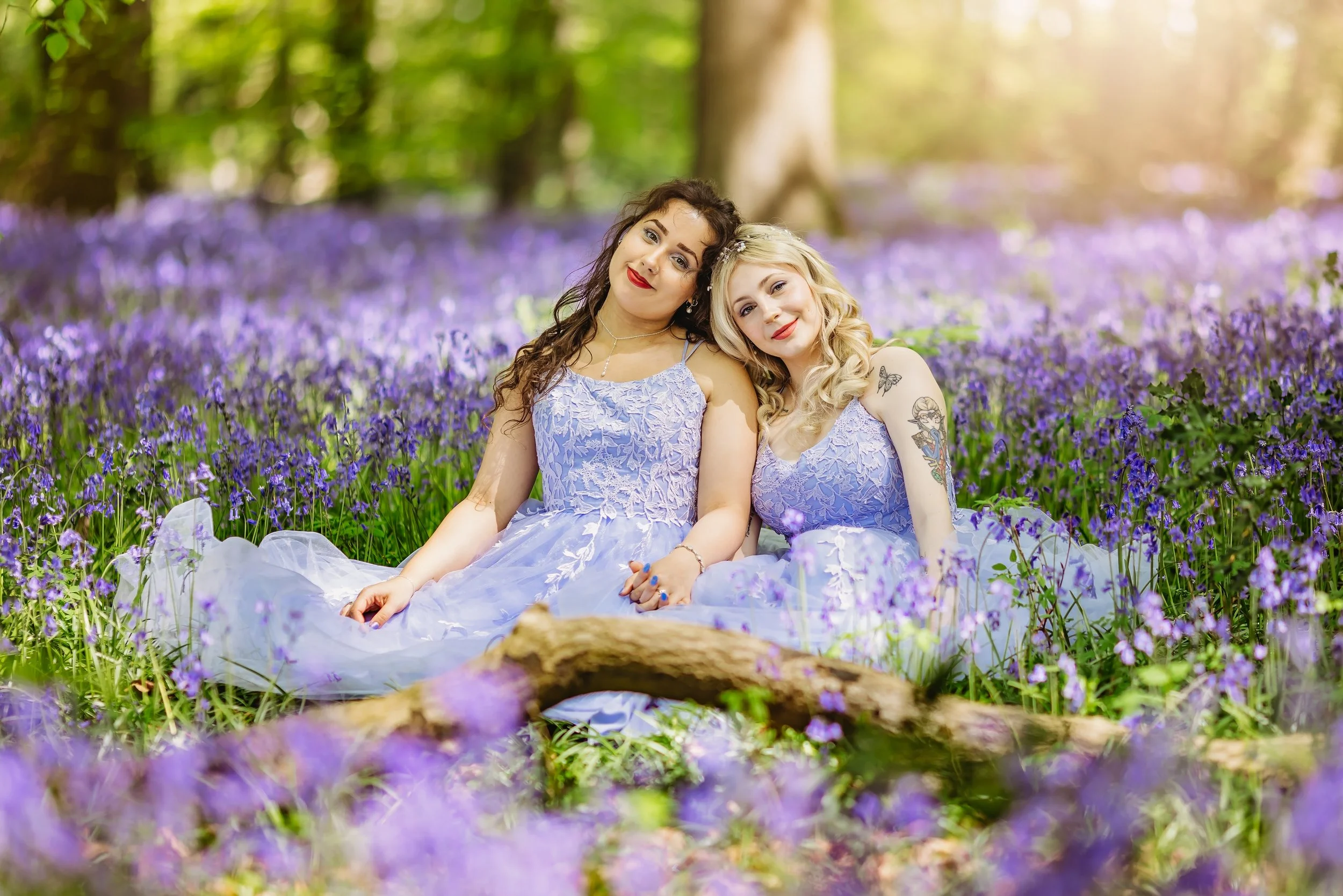 Two women in lavender dresses sitting in a field of purple flowers with trees in the background.