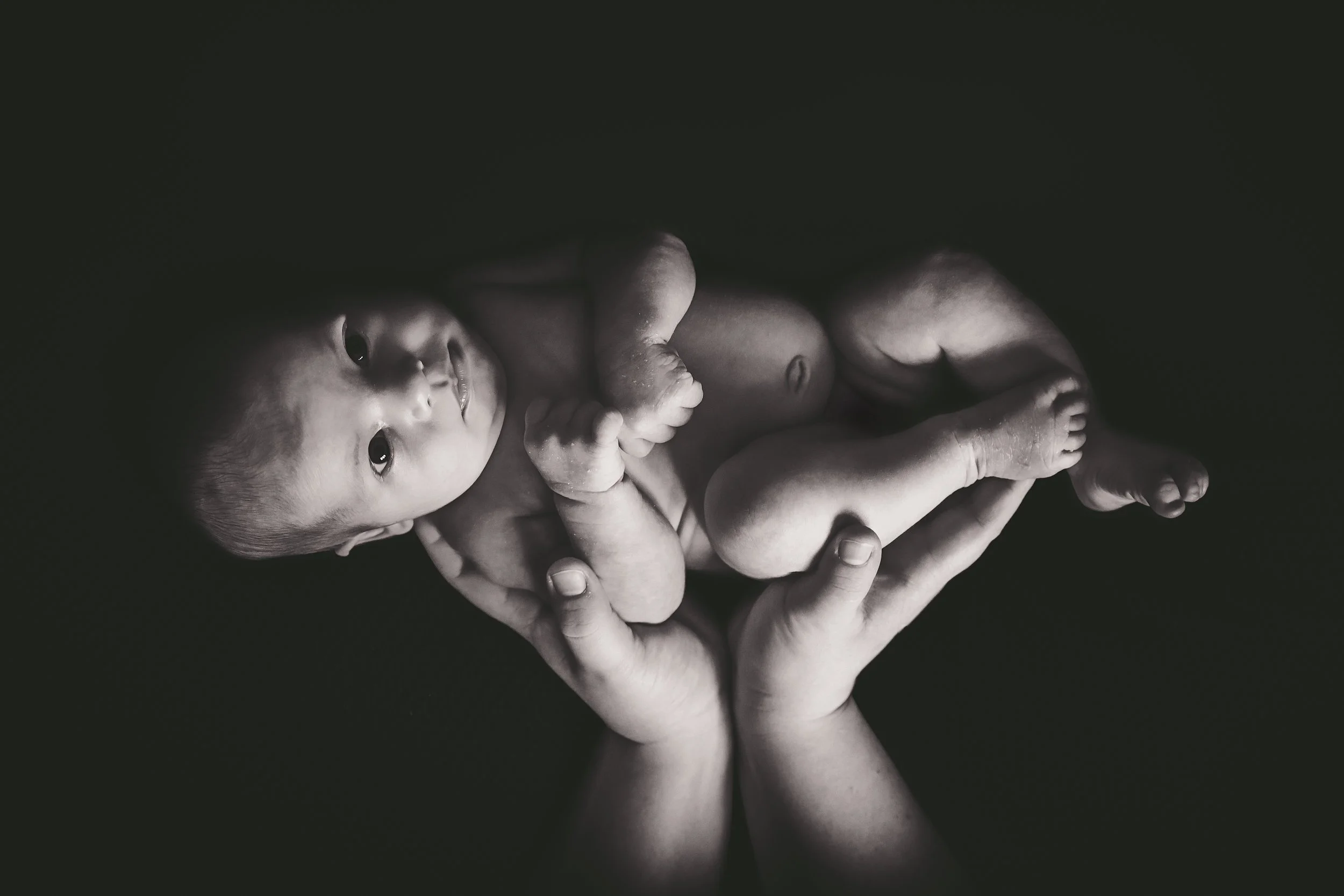 Black and white photo of a newborn baby held gently in adult hands, lying on their side, looking at the camera.