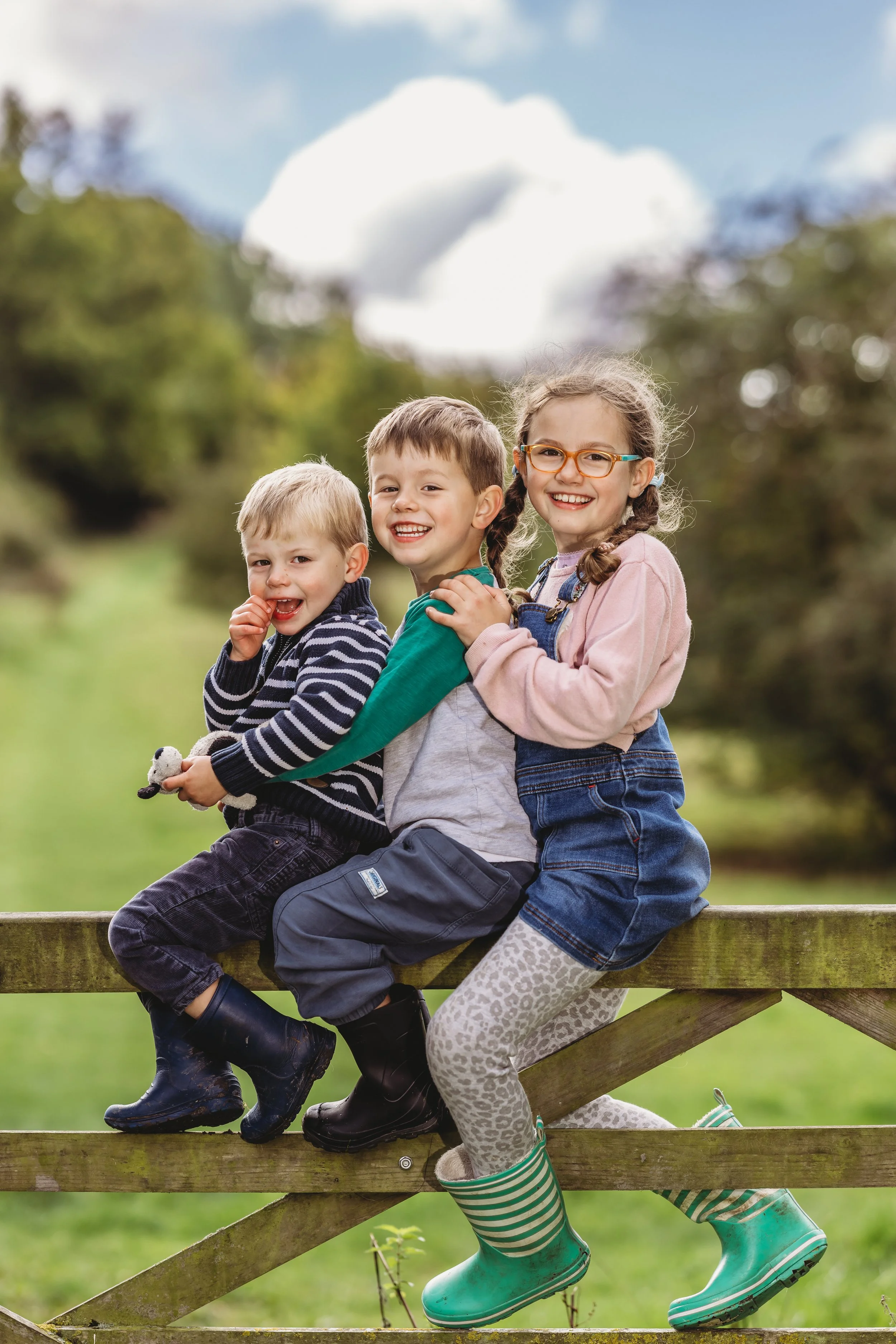 Three children sitting on a wooden fence outdoors, smiling and enjoying a sunny day.