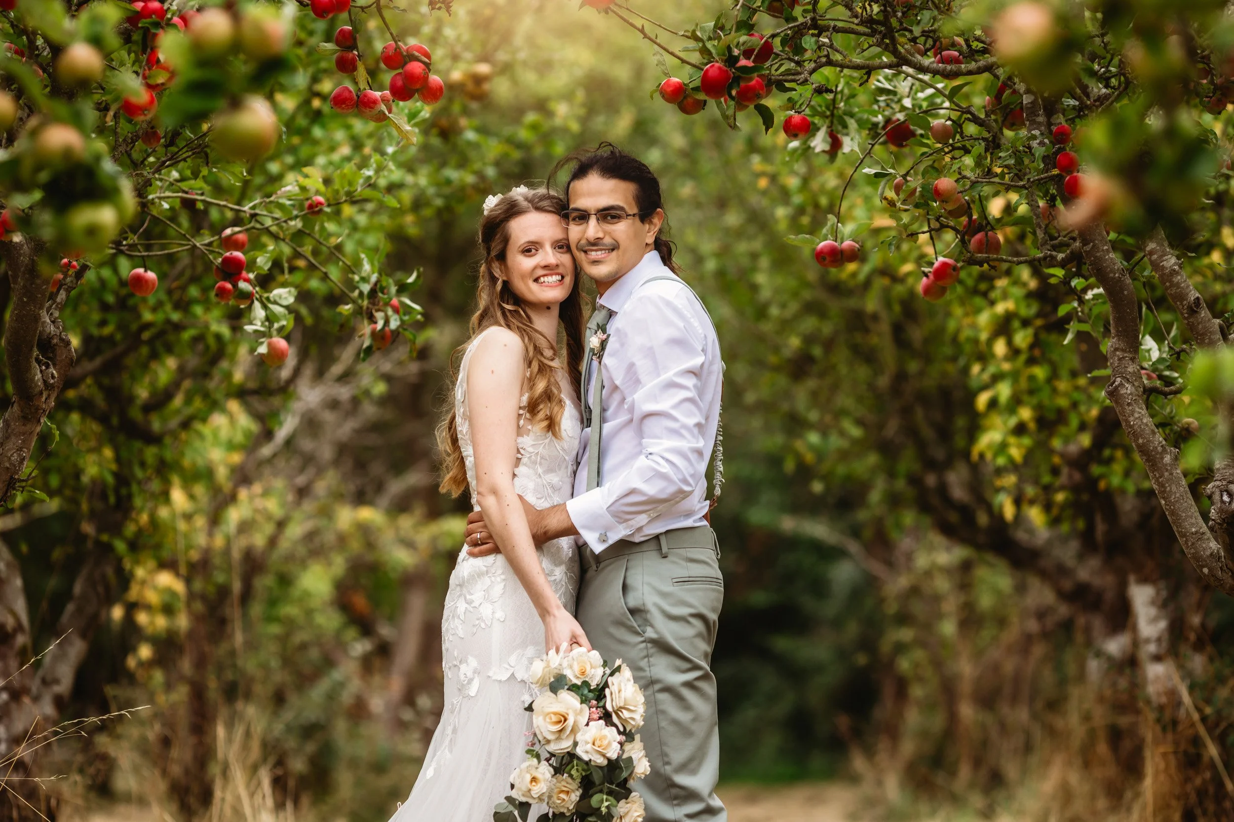 A newlywed couple stands close together in an apple orchard, smiling. The bride holds a bouquet of white flowers, and the groom wears a white shirt and gray pants. Apple trees with red and green apples surround them.