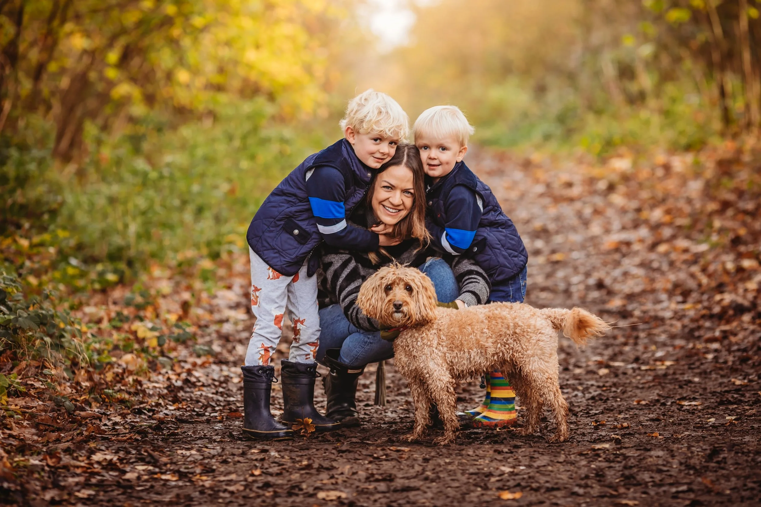 A woman with two young children and a dog on a forest trail during autumn, surrounded by fallen leaves and autumn-colored trees.