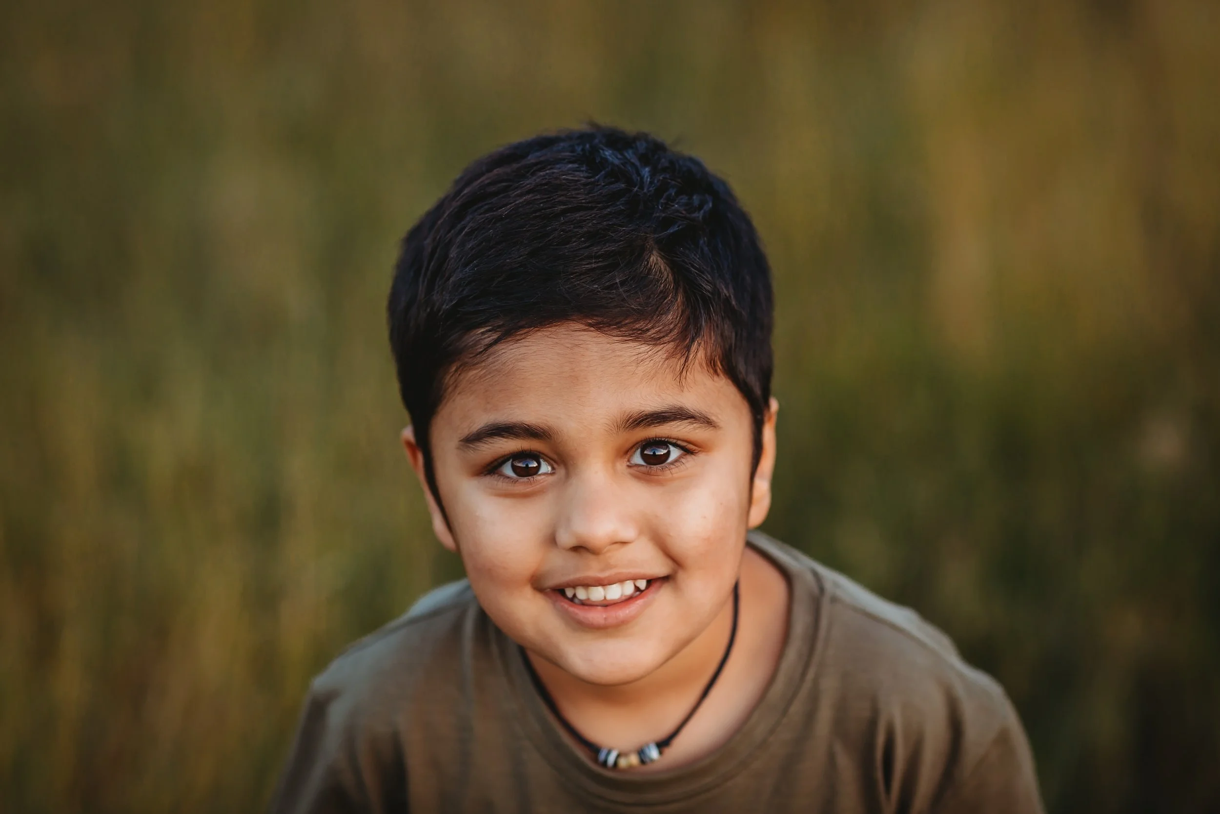 A smiling young boy with dark hair and a necklace outdoors with a blurred green background.