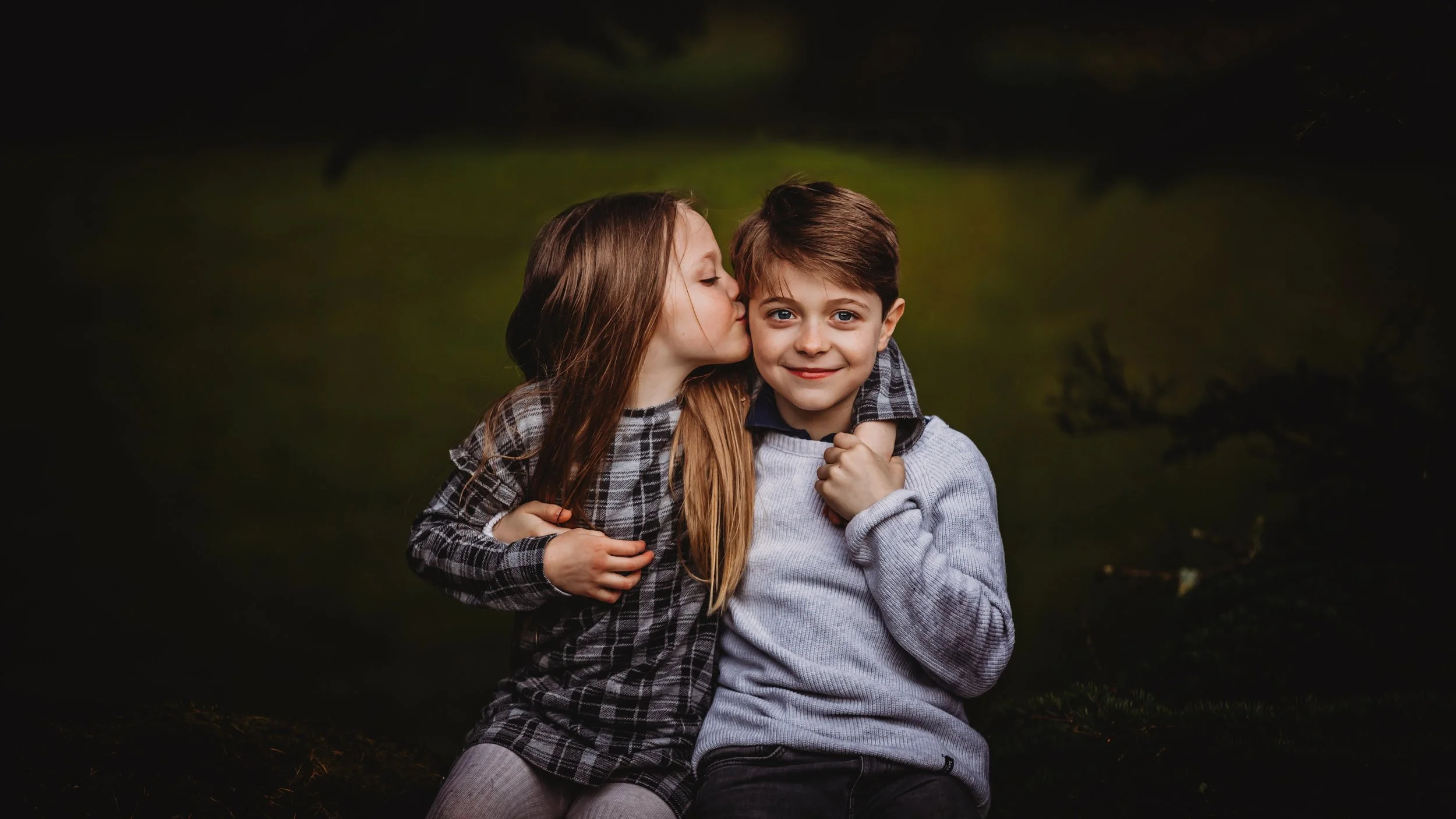 A young girl with long brown hair is giving a kiss on the cheek to a young boy with short brown hair, who is smiling. They are outdoors, dressed in casual clothes, with dark blurred background.
