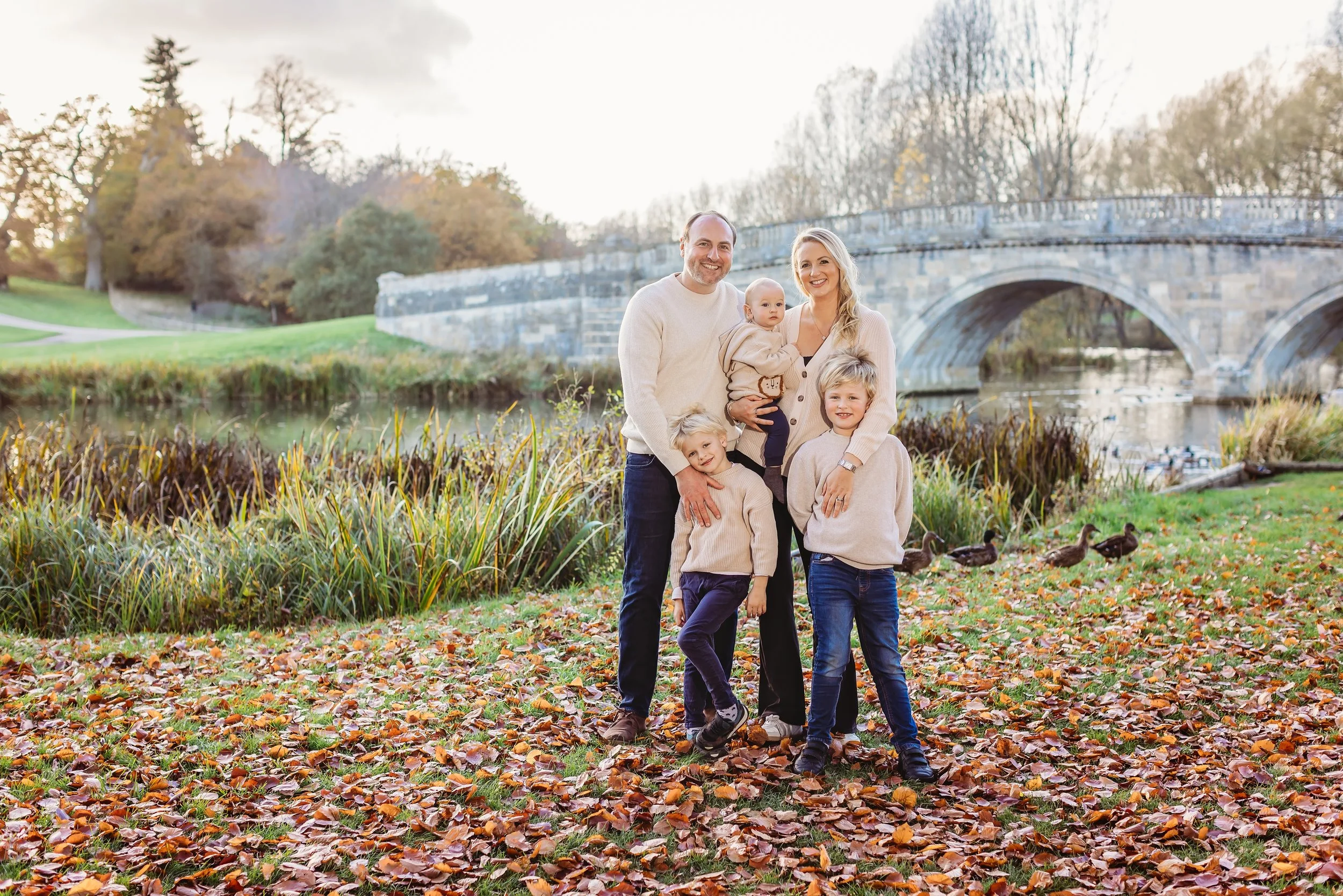 A family of six standing outdoors near a pond with ducks, autumn leaves on the ground, and an arched stone bridge in the background.