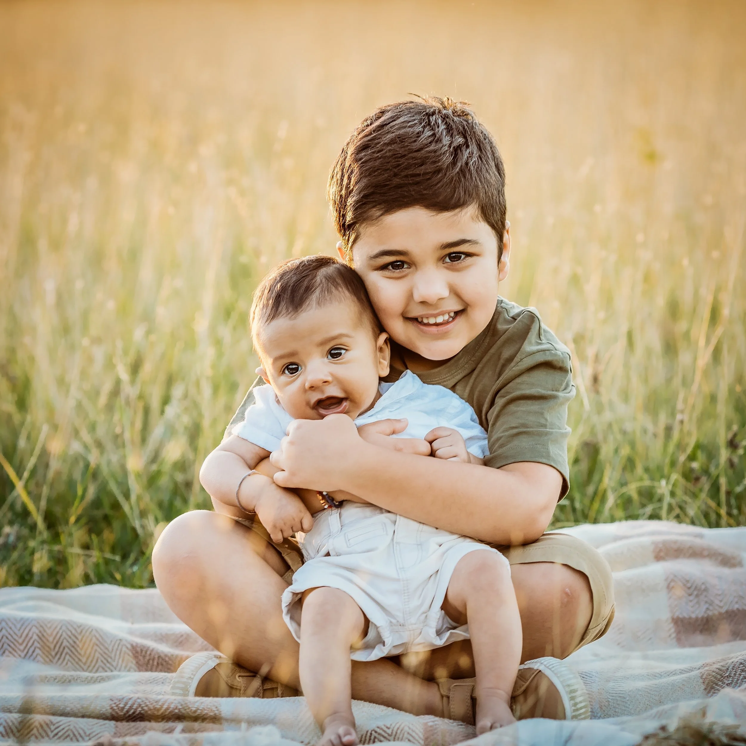 Two young boys sitting outdoors on a blanket in a grassy field, smiling and hugging each other. The older boy has dark hair and is wearing a green shirt, while the younger boy has short dark hair and is wearing a white shirt.