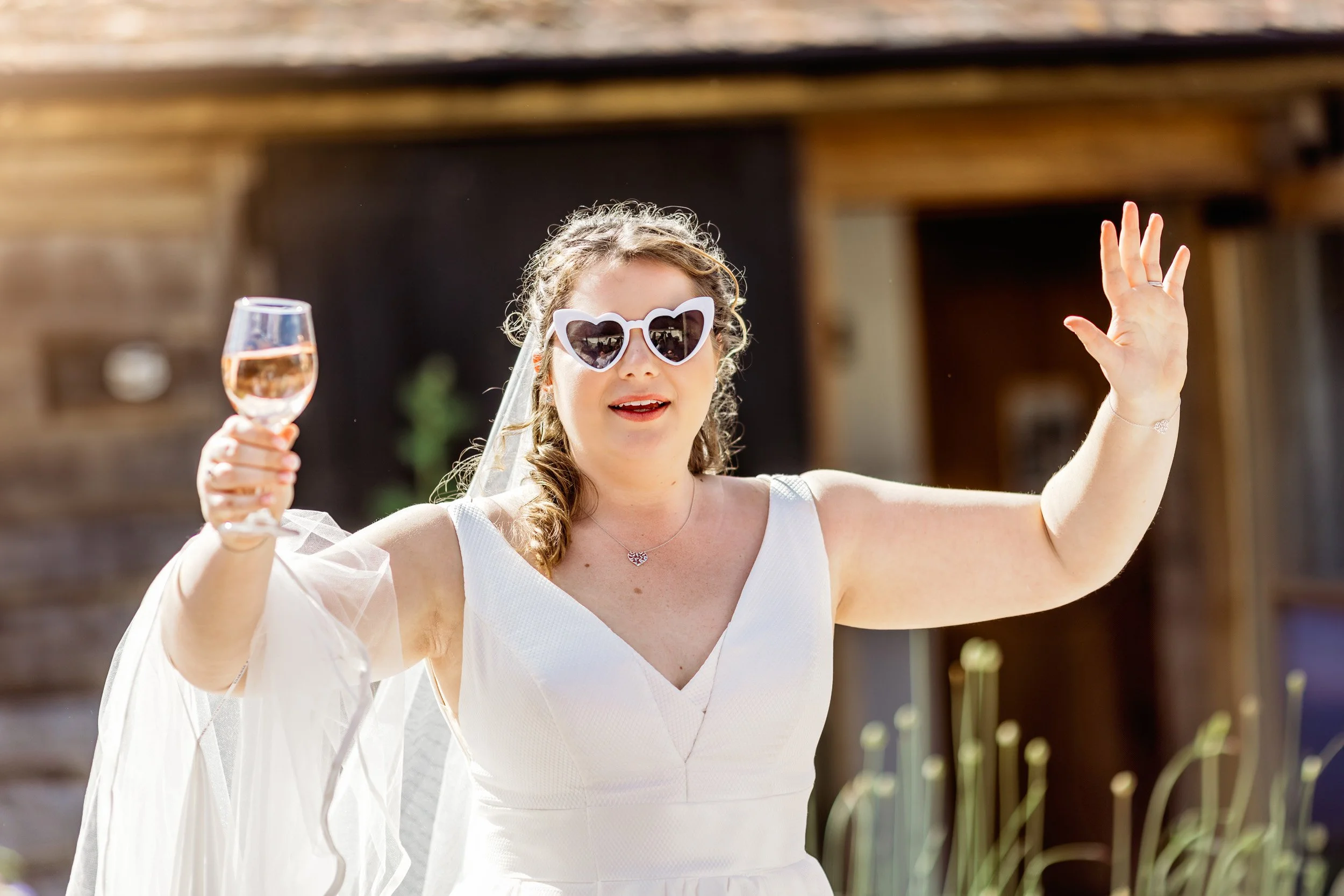 A woman wearing white heart-shaped sunglasses and a white dress, smiling and waving while holding a glass of rosé wine in an outdoor setting.