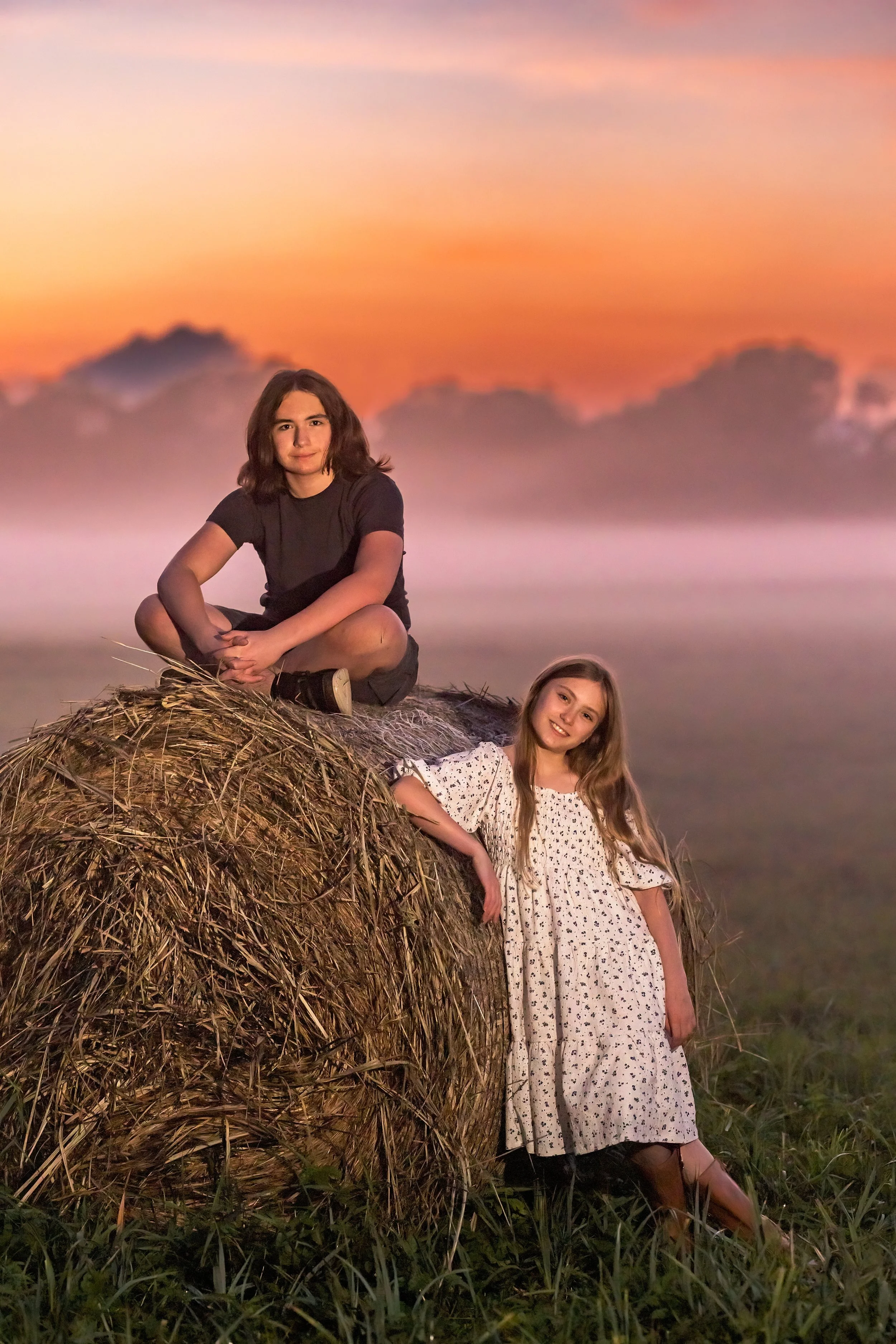 Two young women pose on a haystack in a field during sunset with a colorful sky and distant clouds.