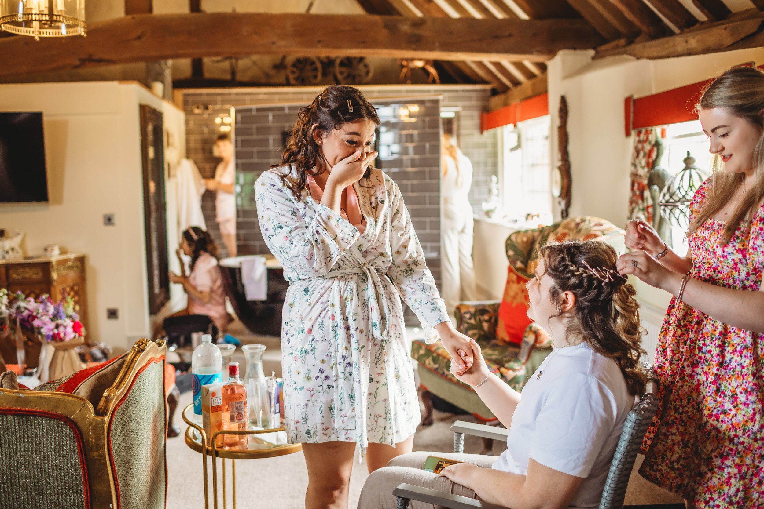 Women with one seated getting her hair styled, while another behind her styles her hair, in a cozy, decorated room with other women preparing in the background.
