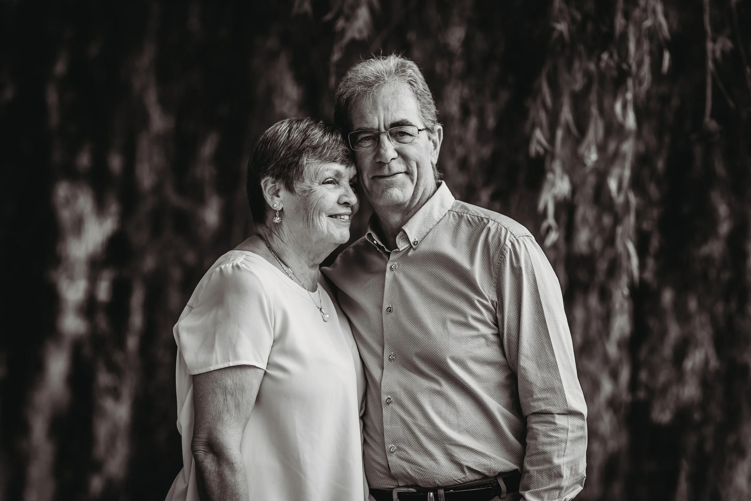 A black-and-white photo of an older woman and man standing close together outdoors, smiling and looking content, with a blurred natural background.