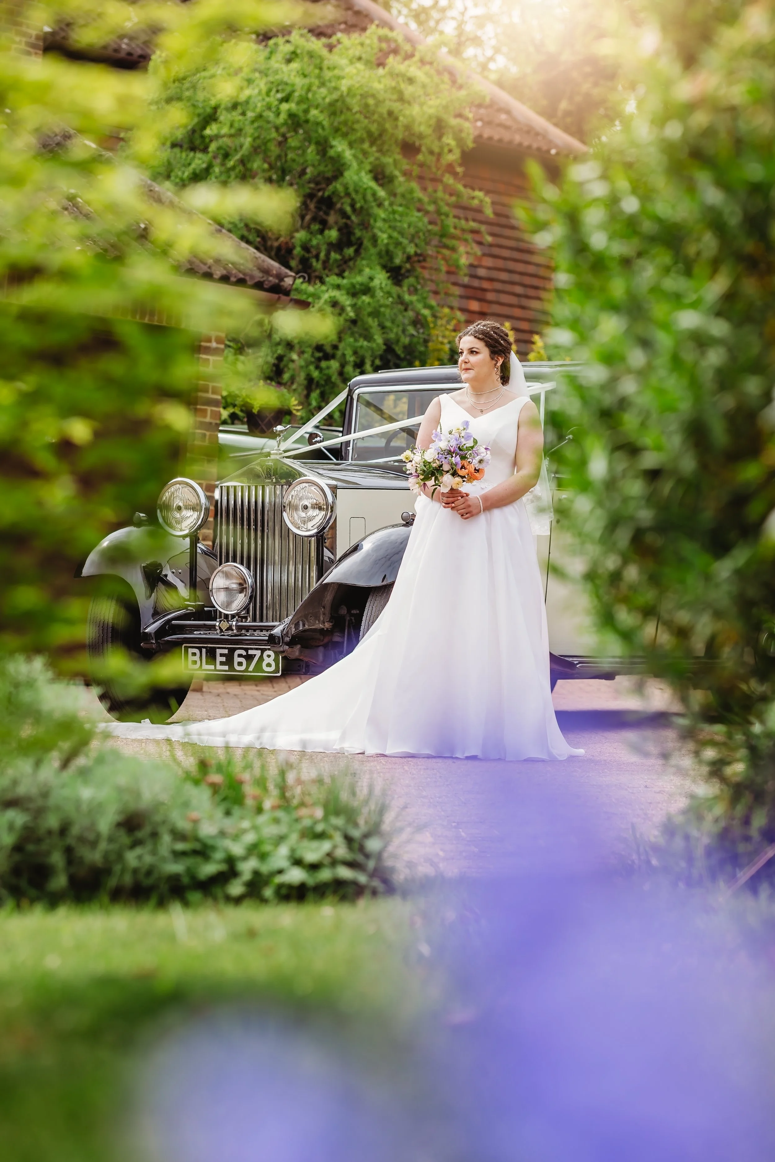 A bride in a white wedding dress with a bouquet of flowers stands next to a vintage black and white car, framed by green bushes and trees.