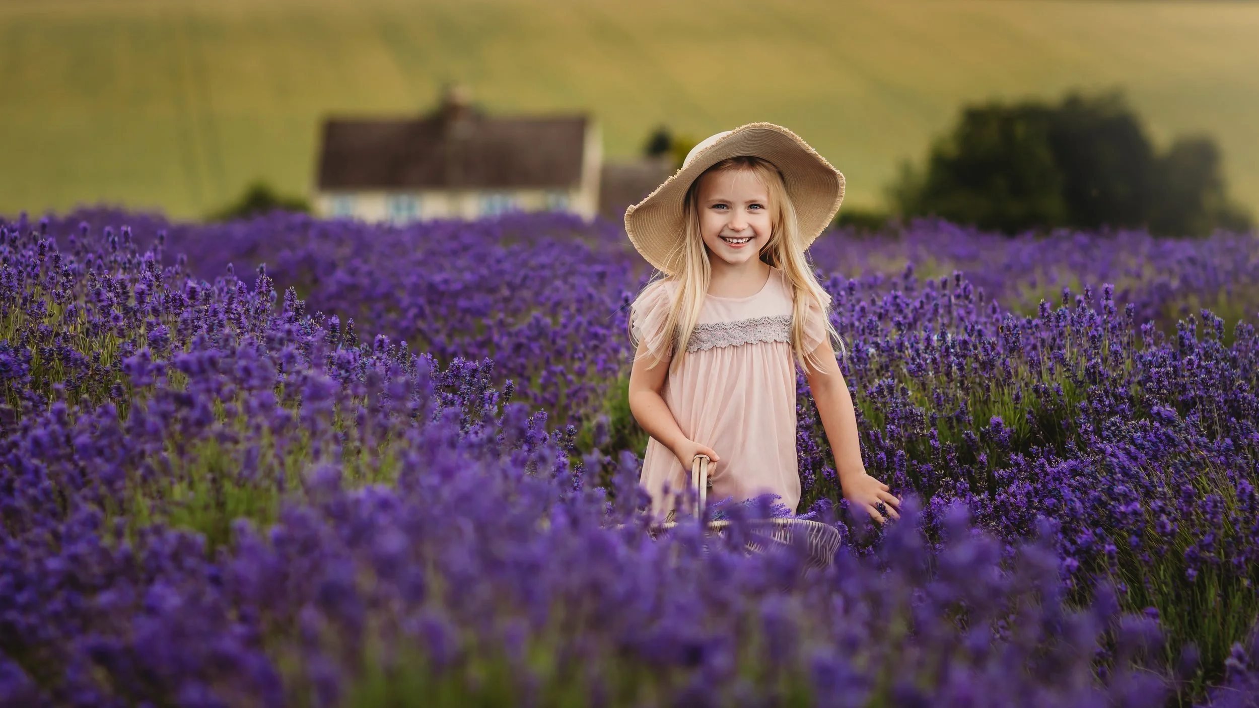 A young girl with long blonde hair wearing a pink dress and a large straw hat smiling in a field of purple lavender flowers with a house in the background.