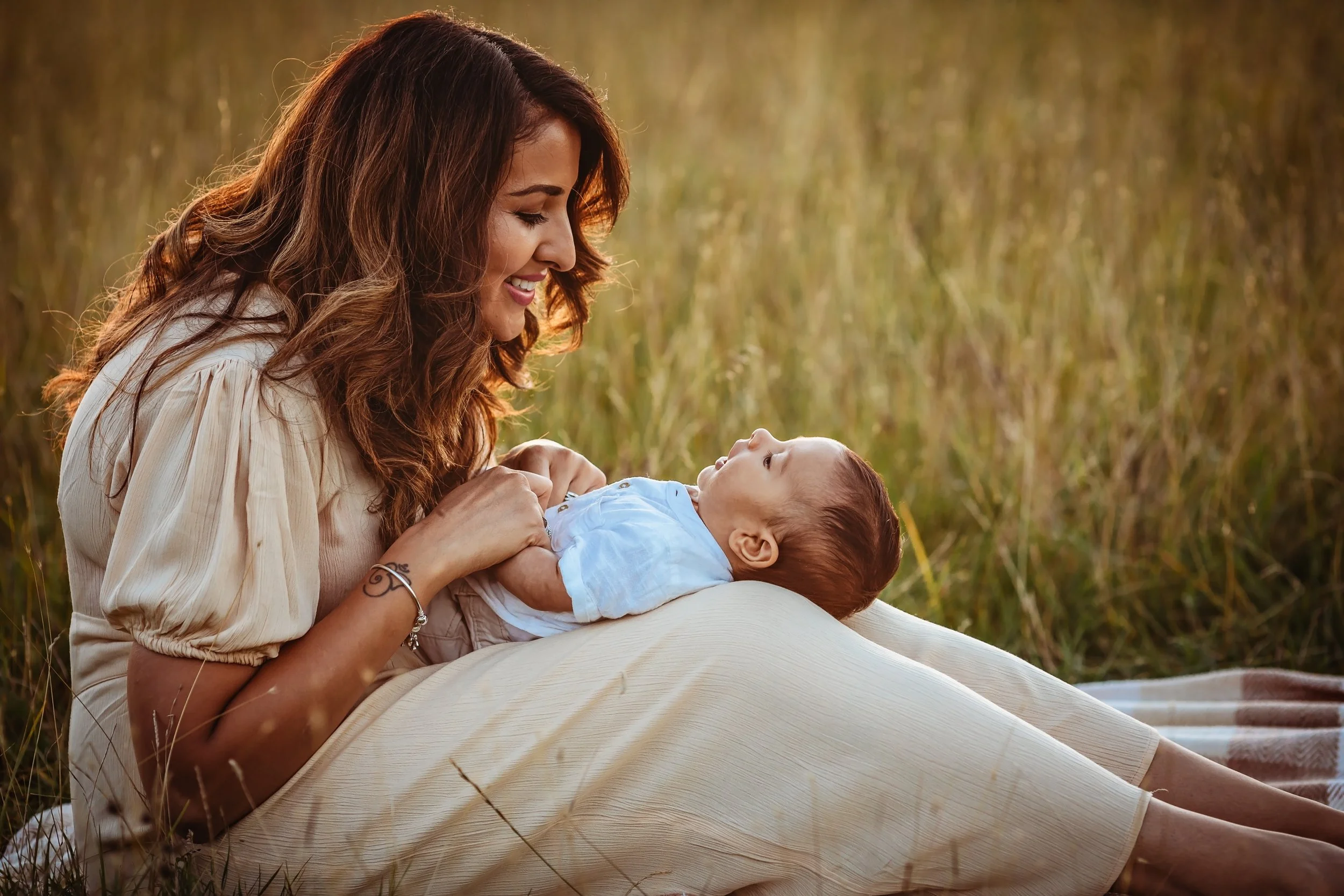 A woman with long brown hair holding a baby boy in a field of tall grass at sunset, both smiling and enjoying each other's company.