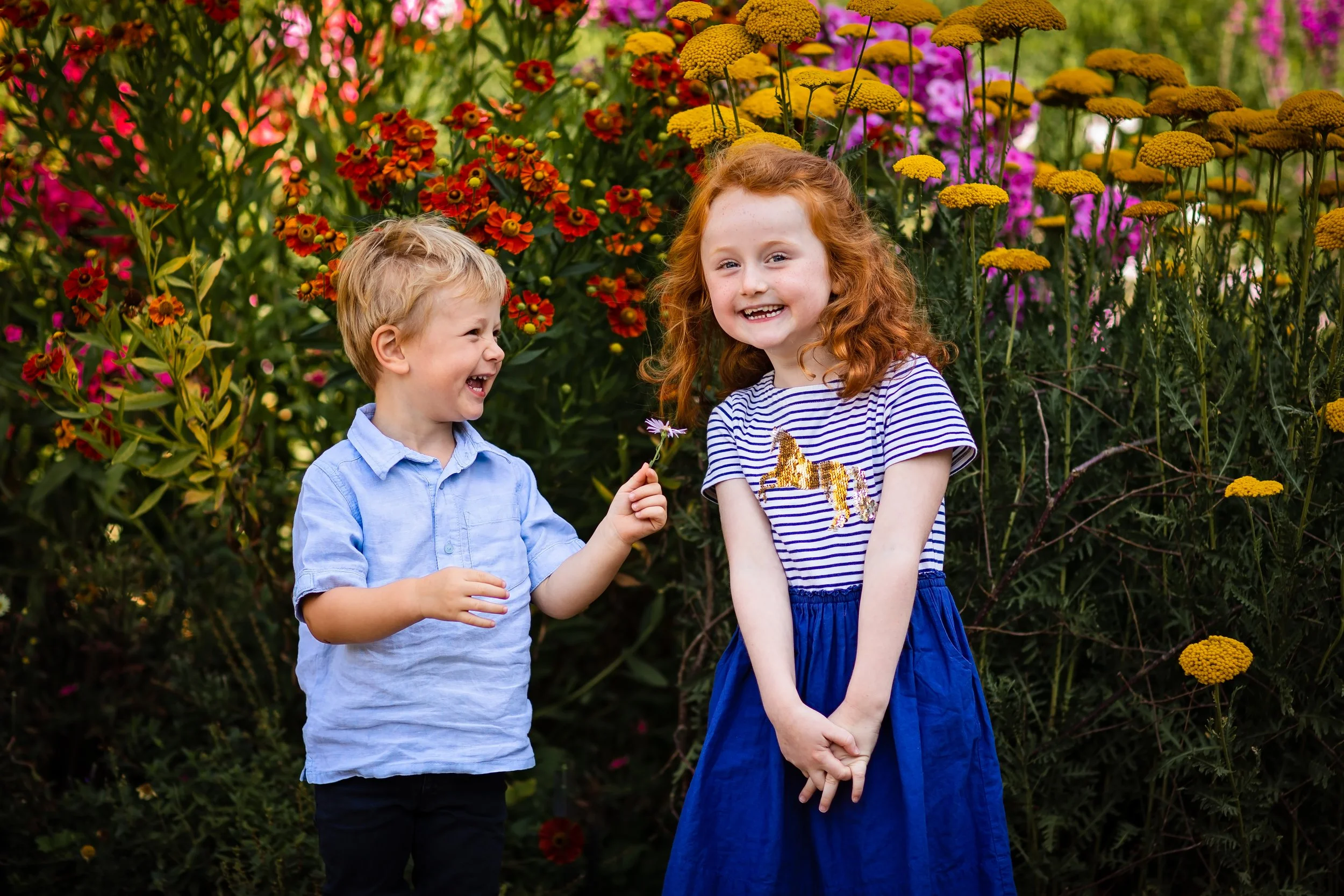 Two children, a boy and a girl, smiling and interacting in a colorful flower garden. The boy is handing a flower to the girl, both are happy and surrounded by vibrant flowers.