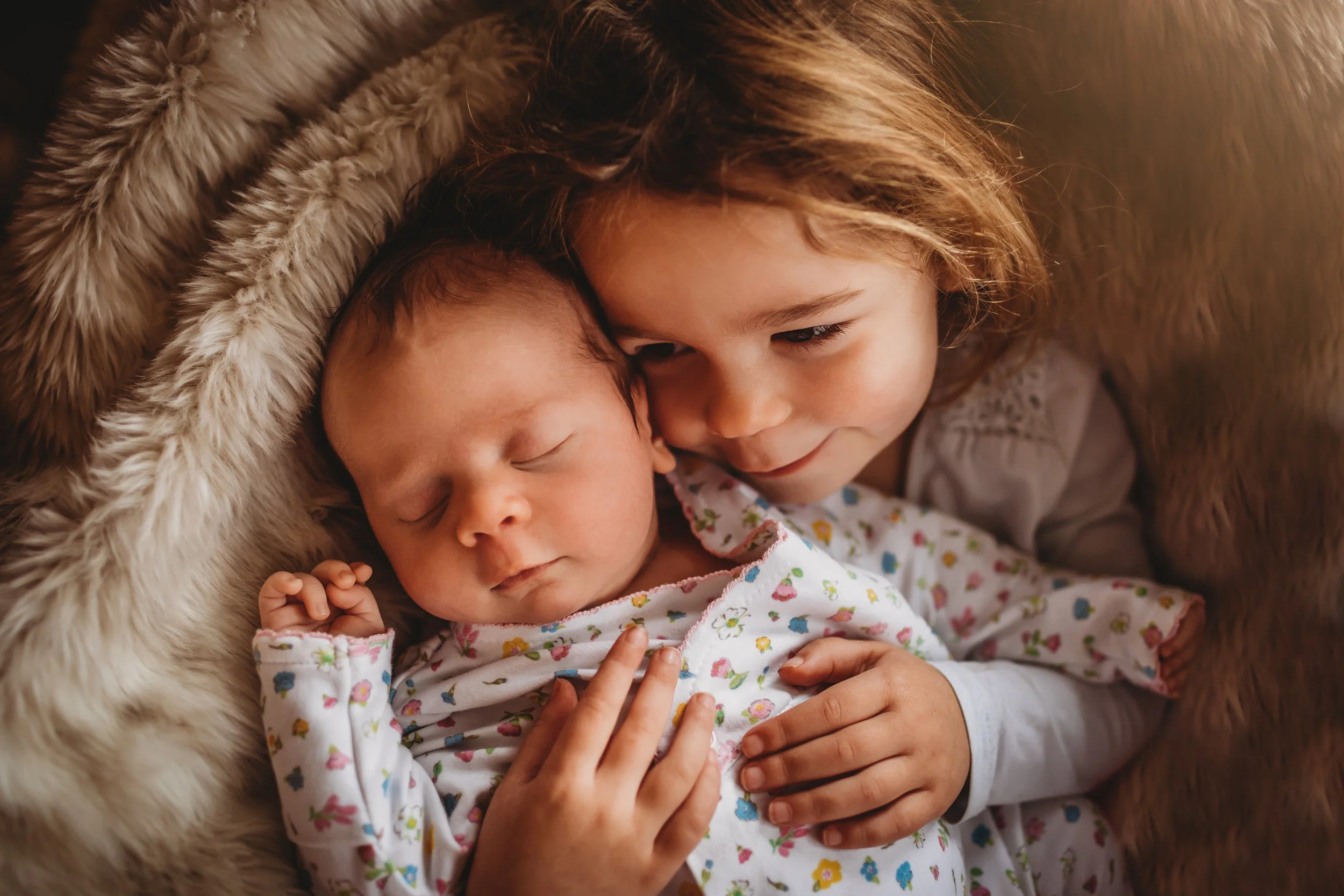 A young girl cuddling a sleeping baby wrapped in a floral blanket, both lying on a cozy fur blanket.