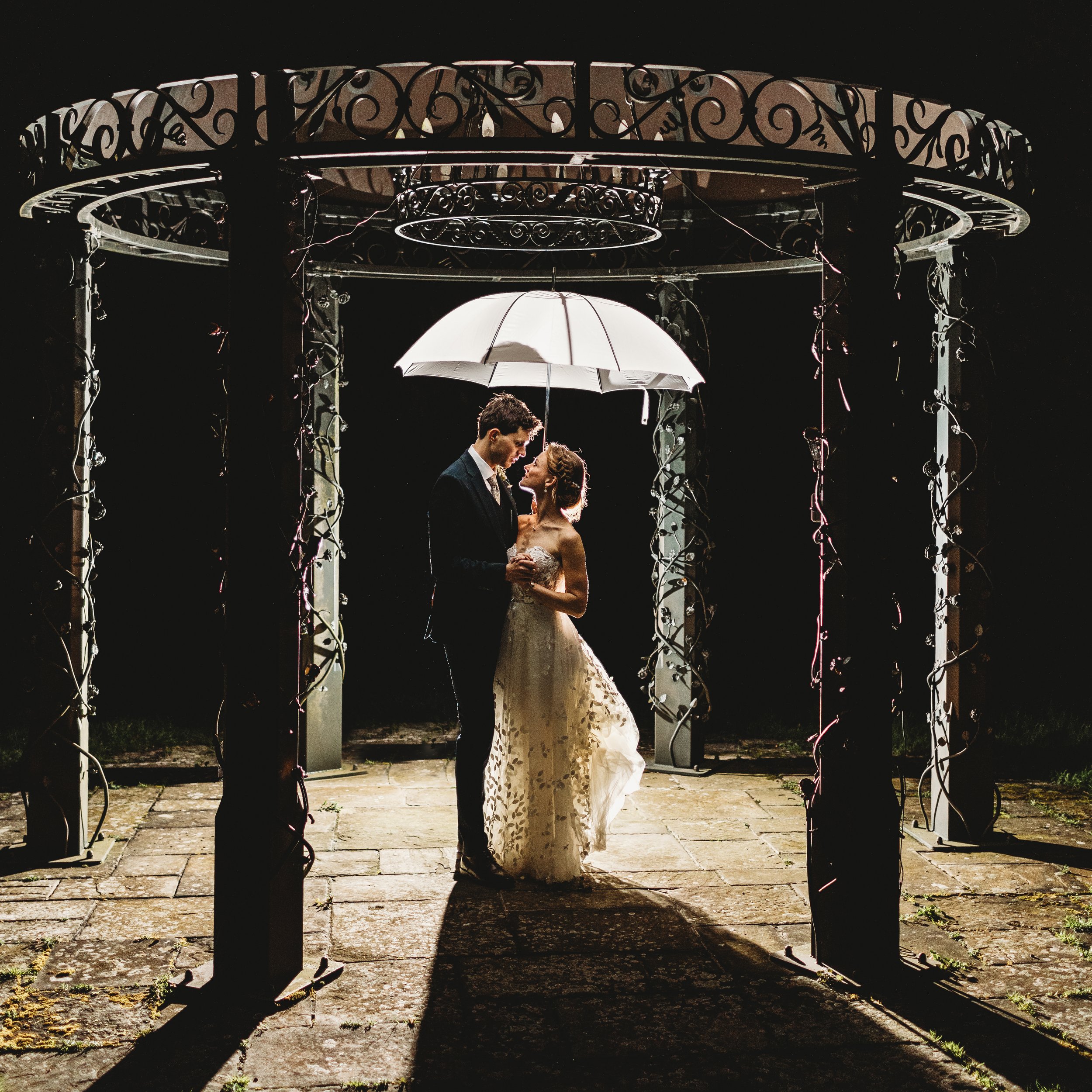 A couple in wedding attire standing under a white umbrella during a nighttime outdoor event, framed by an ornate metal arbor.