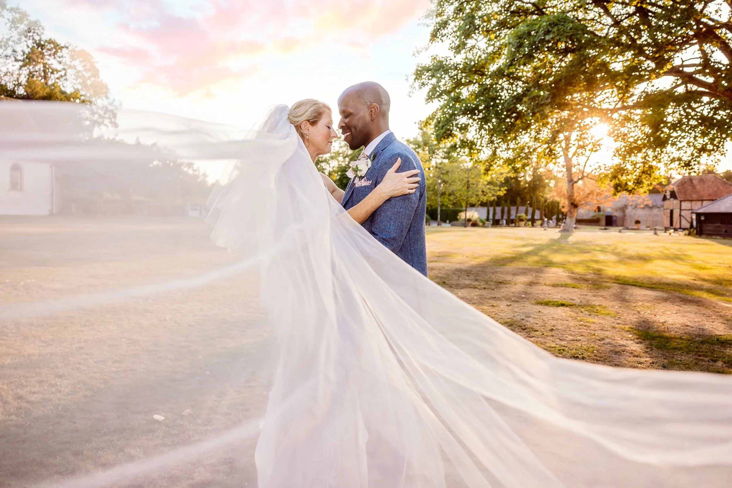 A wedding couple holding each other in a park during sunset, with the bride's veil flowing in the wind.