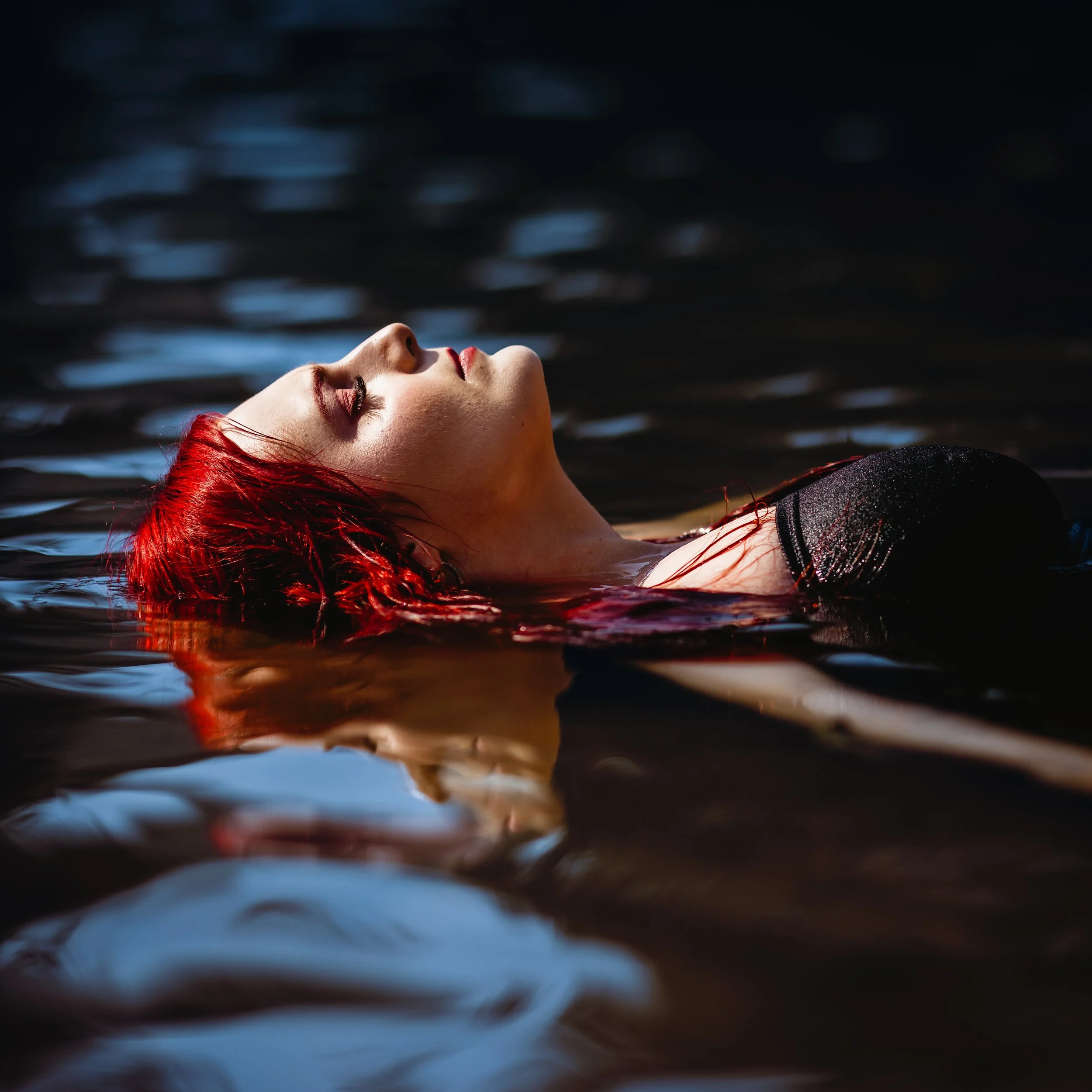 A woman with red hair floating on her back in dark water with her eyes closed.