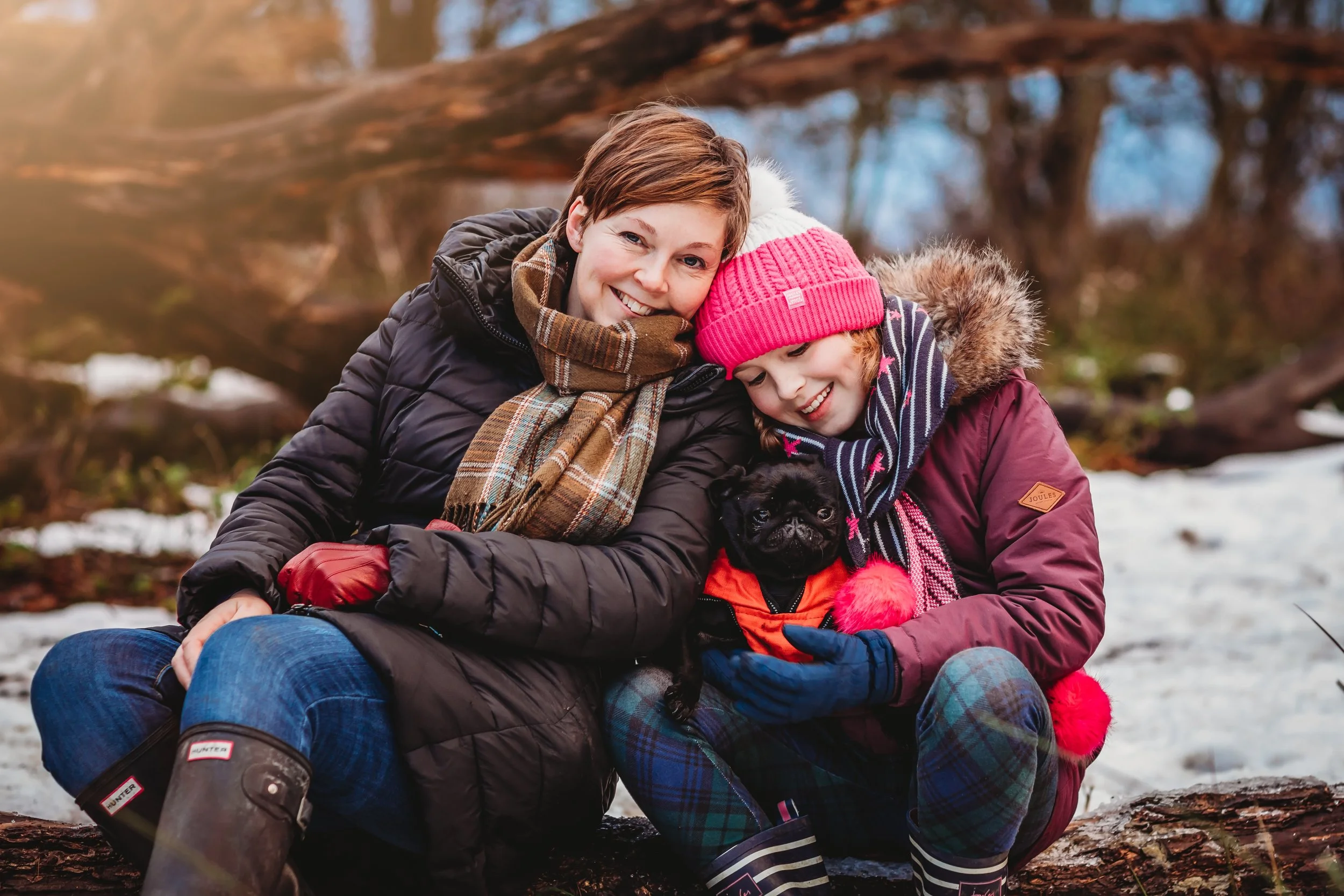 A woman and a girl sitting outdoors on a log, smiling and cuddling a small black dog wearing an orange jacket. The woman is wearing a black jacket and a plaid scarf, the girl is wearing a pink beanie, a maroon jacket with fur hood, and striped pants.