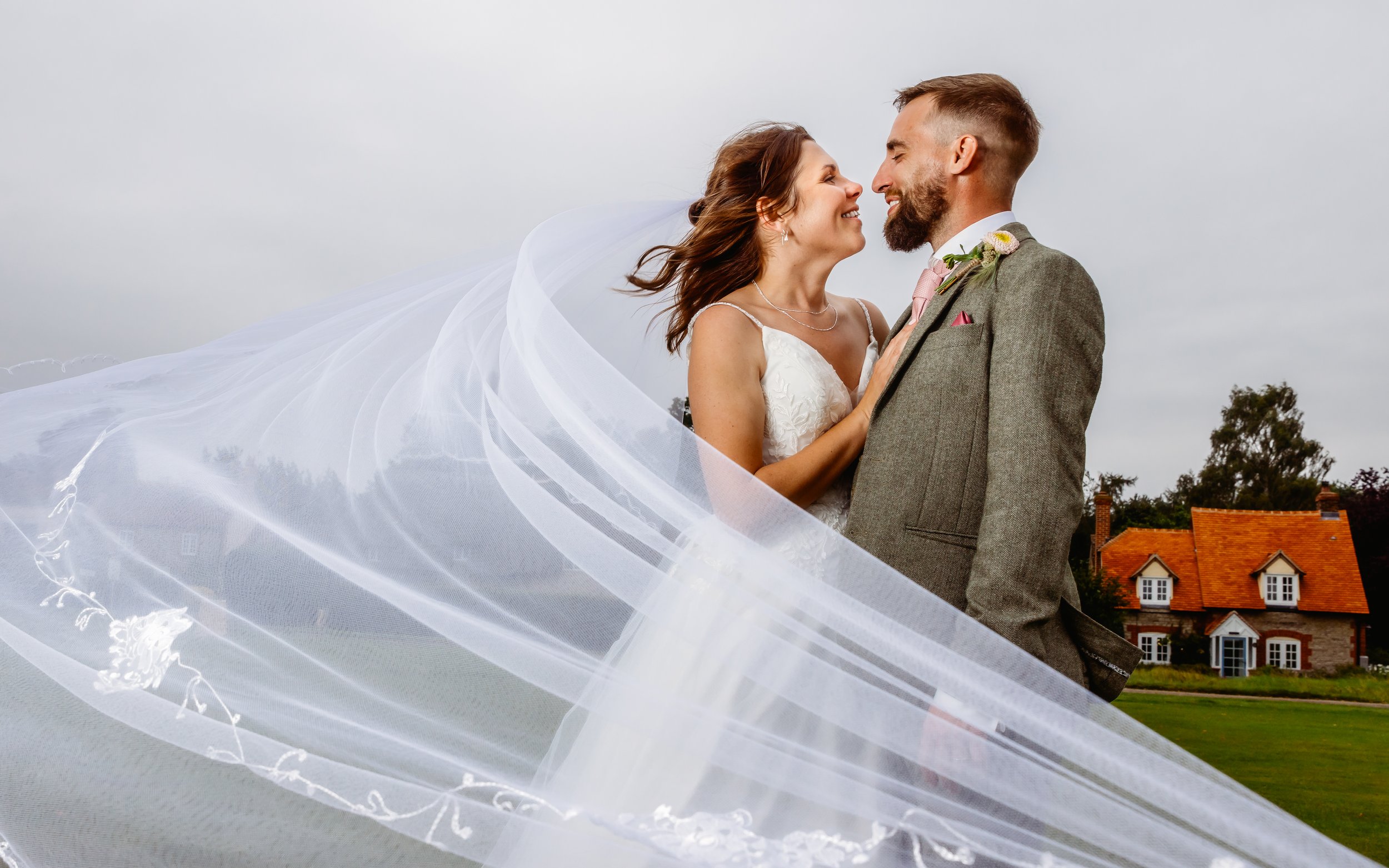 A bride and groom stand close, smiling at each other on their wedding day outdoors with a large white veil flowing around them and a house in the background.