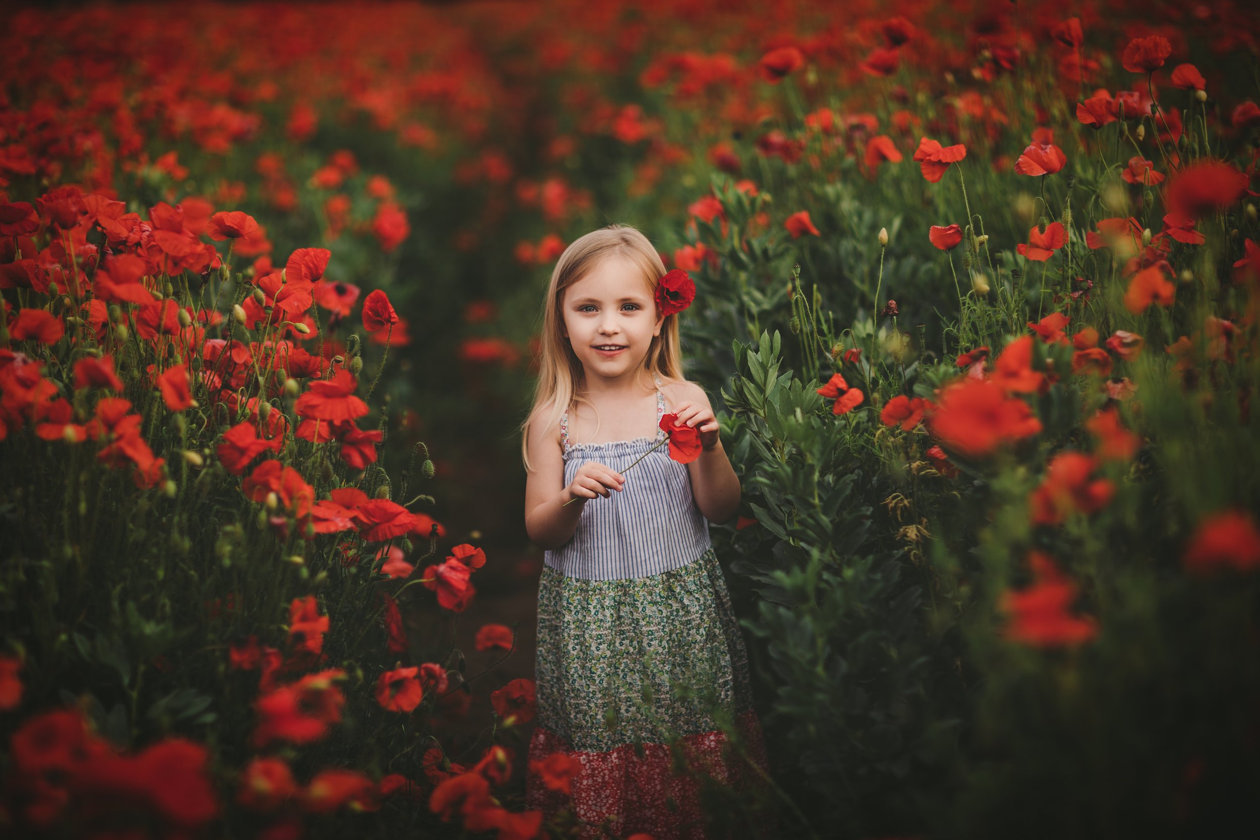 A young girl with blonde hair, wearing a striped top and floral skirt, standing in a field of red flowers, holding a flower and smiling.