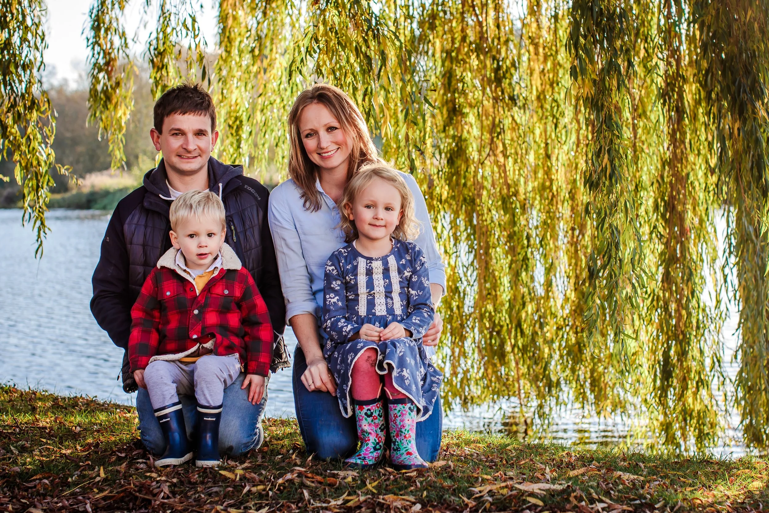 Family of five outdoors near a lake with golden trees in the background, smiling and sitting on fallen leaves.