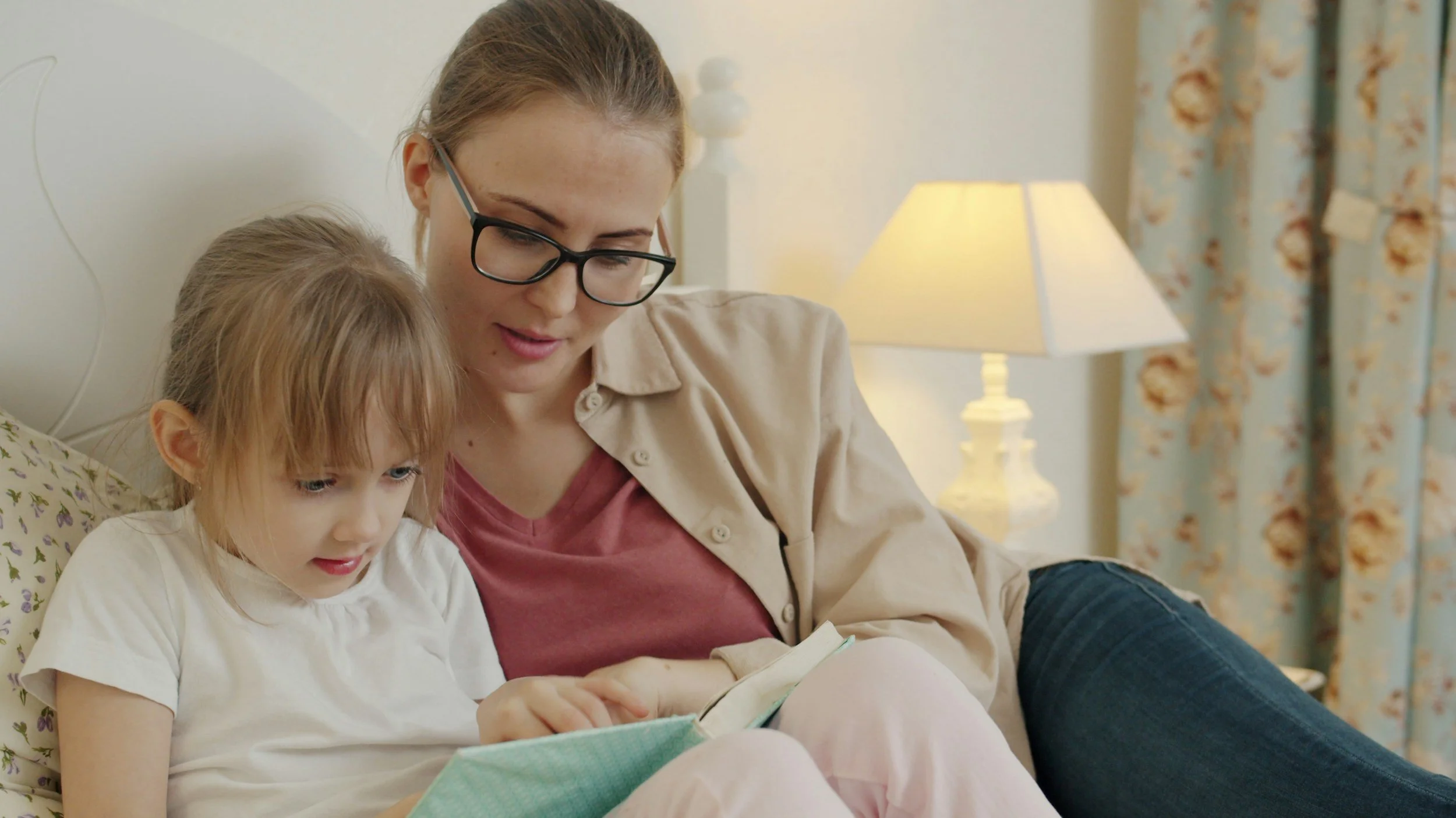 Vrouw en kind lezen samen een boek op de bank in een gezellige woonkamer met een lamp en bloemenpatroon op de gordijnen.