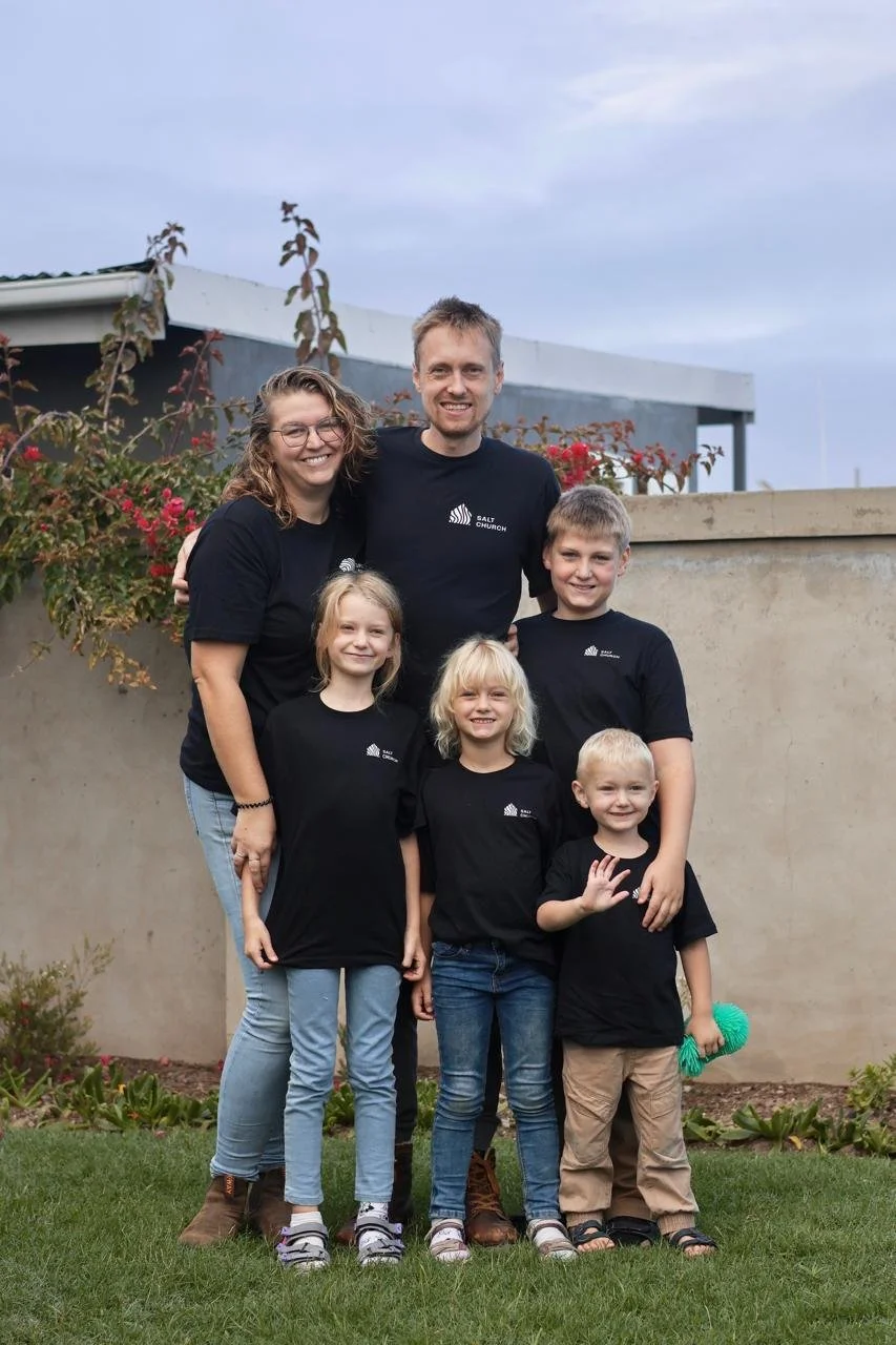 A family of seven standing outdoors in front of a concrete wall and garden, all wearing matching black T-shirts with white logo, smiling for the photo.