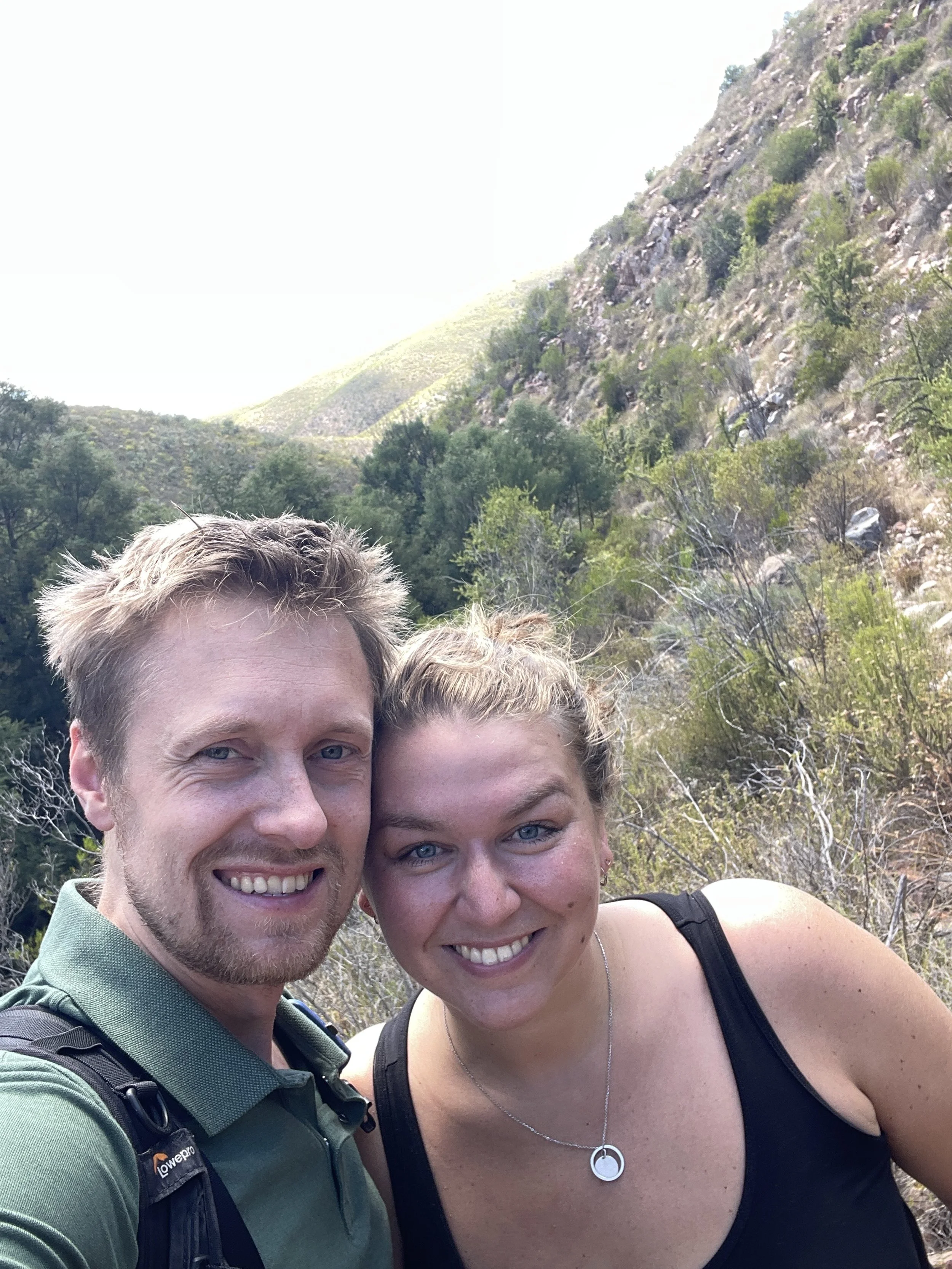 A smiling couple taking a selfie outdoors in a mountainous area with trees and rocky slopes.
