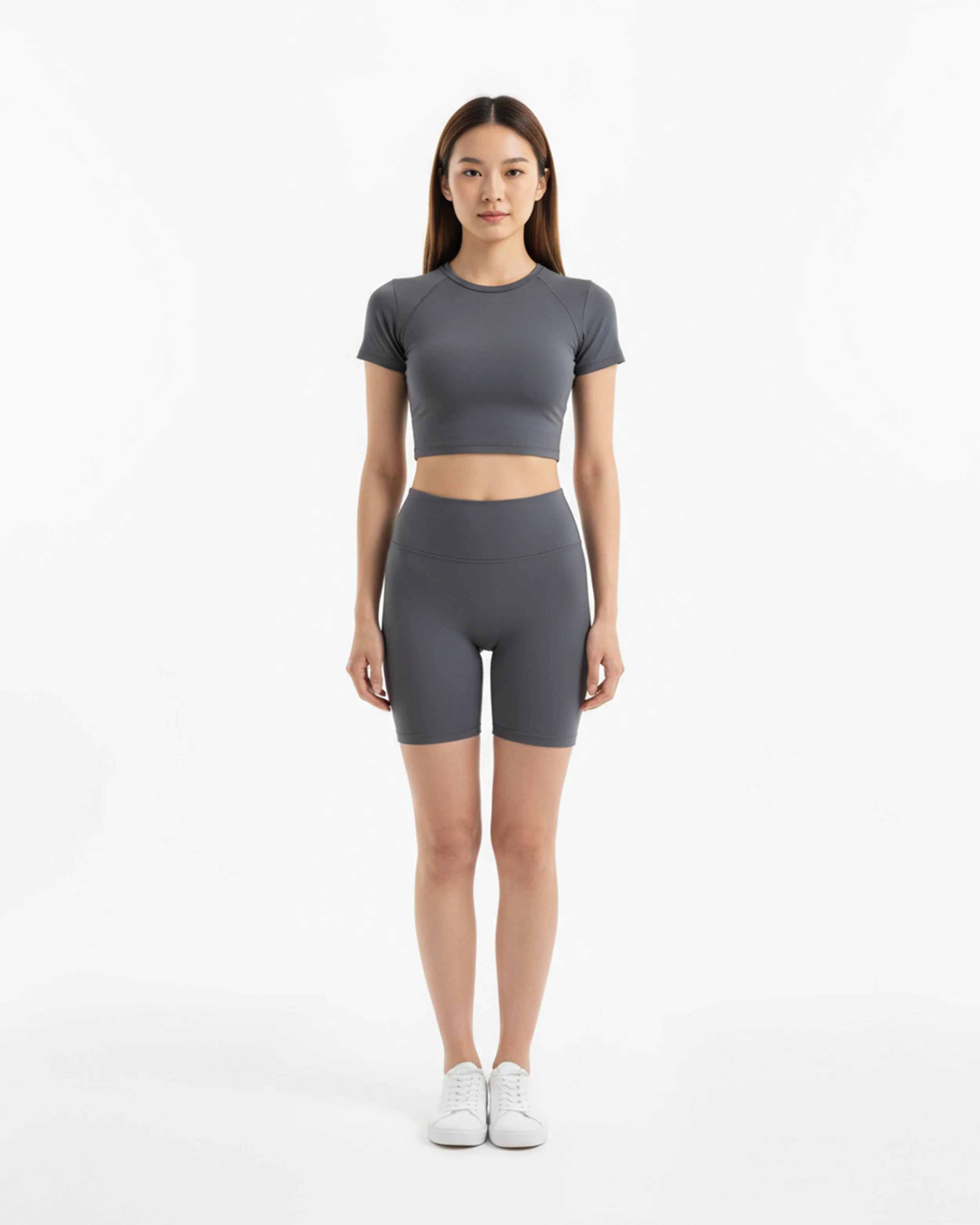 Donna in abbigliamento sportivo grigio, con pantaloncini e maglietta, in piedi su sfondo bianco.